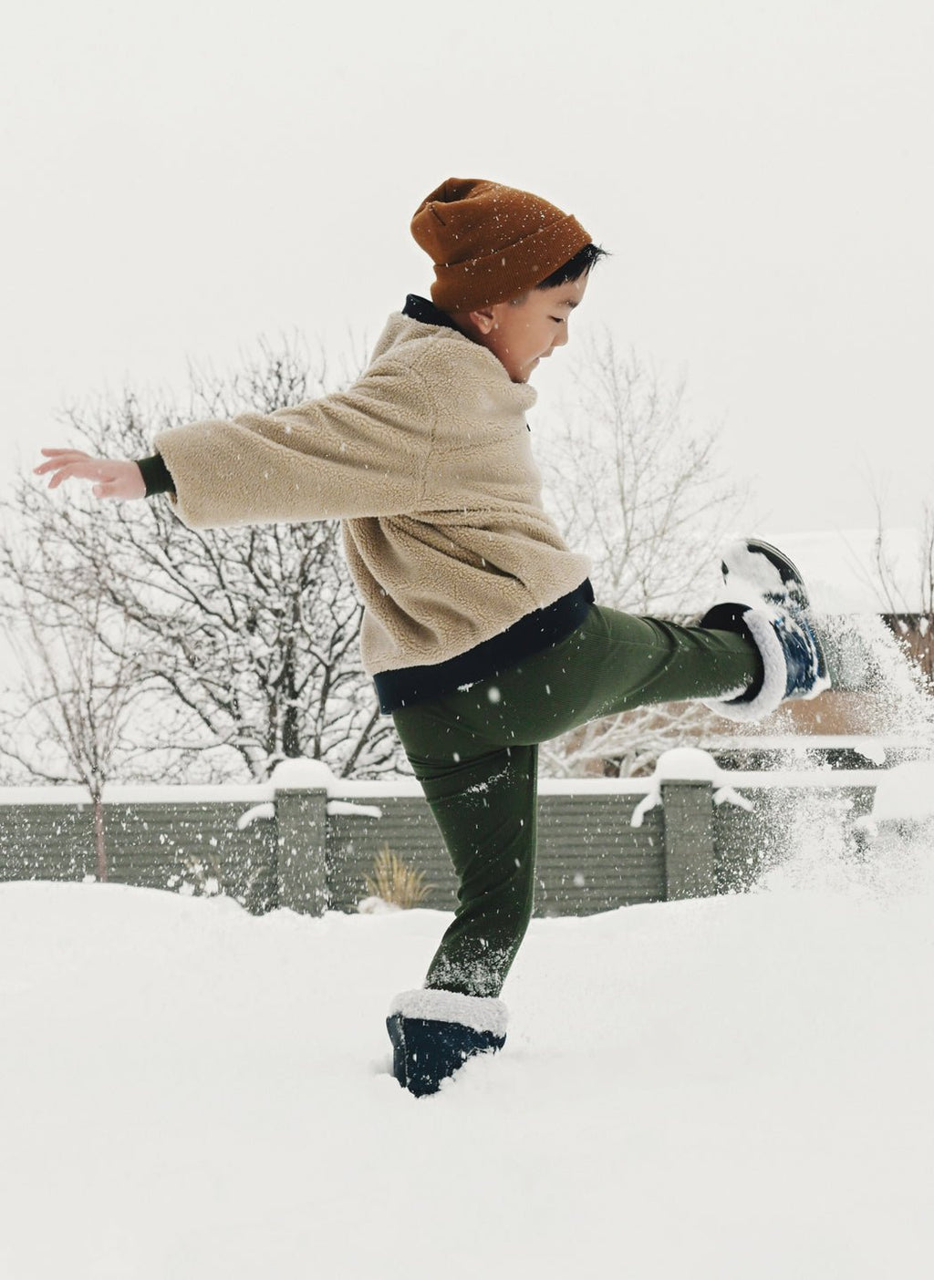 A young child wearing Cozy Earth's Children's Bamboo Waffle Knit Base Layer Pant plays outside, kicking up snow on a wintry day with trees and a fence in the background.|Color:Olive