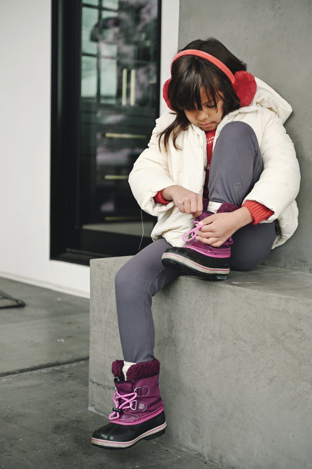 A young girl in Cozy Earth Children's Bamboo Waffle Knit Base Layer Pants sits on a concrete ledge, tying her boot. She also wears a white winter coat, purple boots, gray leggings, and red earmuffs.|Color:Iron