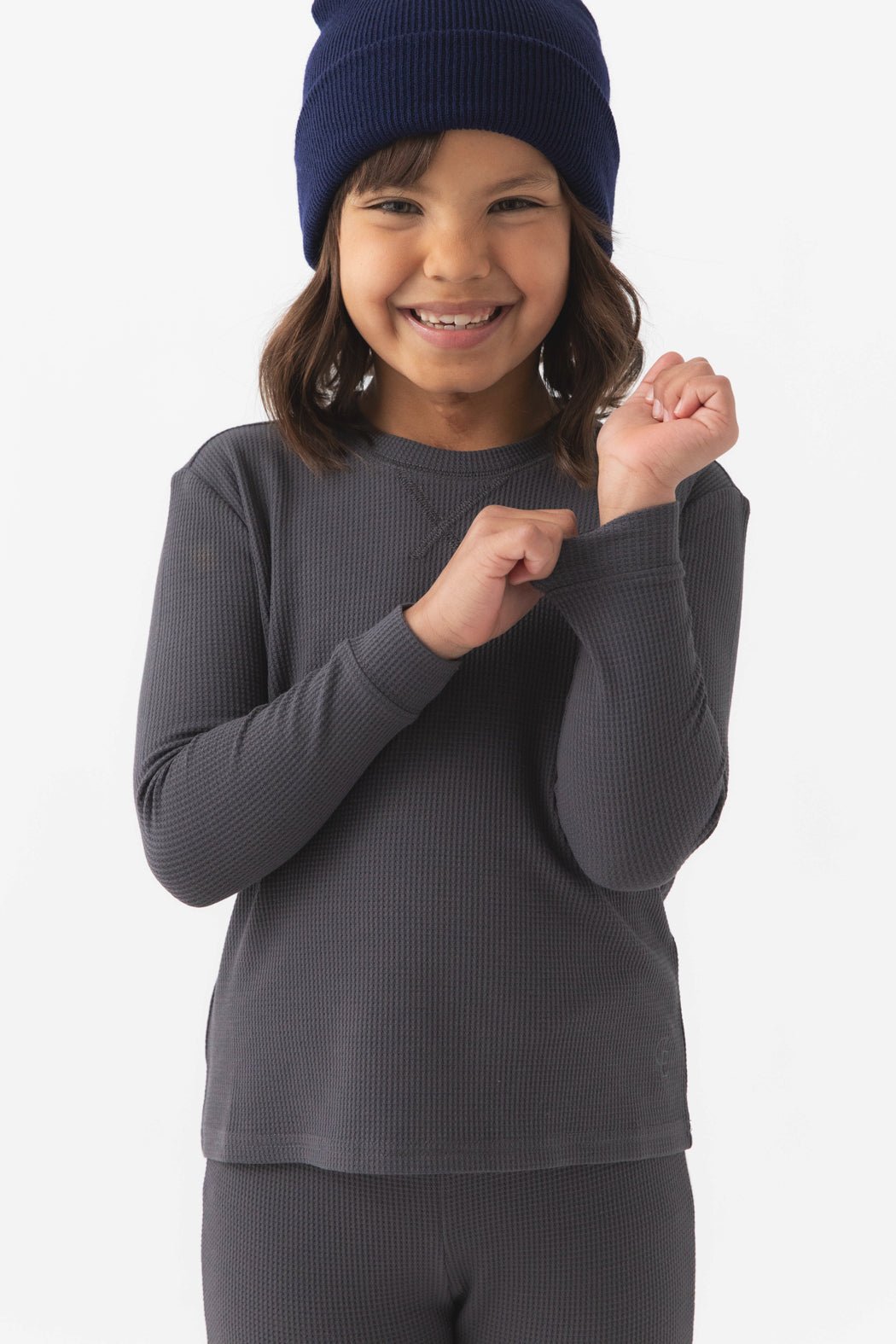 A young child wearing Cozy Earth's Children's Bamboo Waffle Knit Base Layer Long Sleeve in dark gray smiles while holding the sleeve. The beanie is dark blue and the background is plain white.|Color:Iron