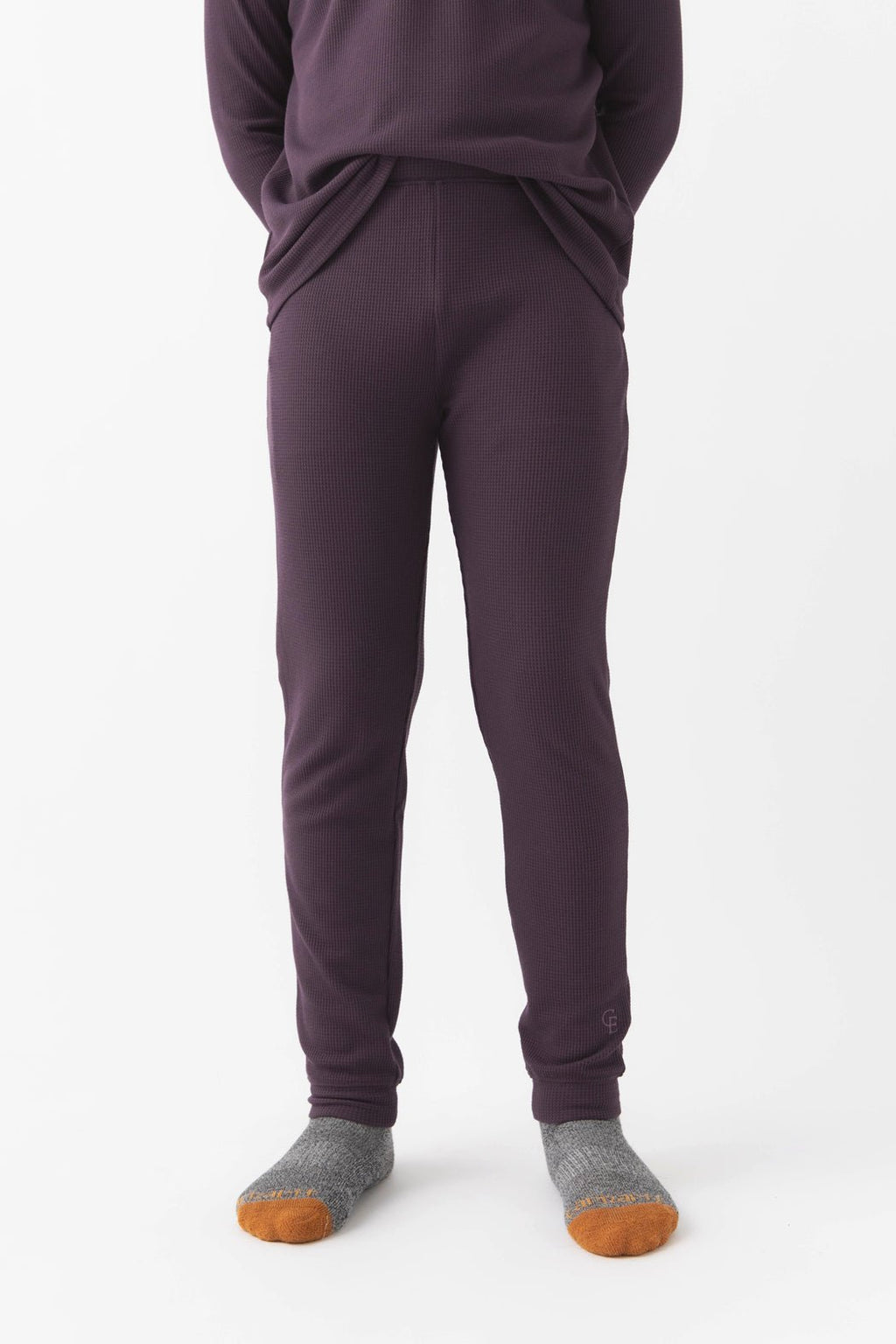 A child stands against a white background wearing Cozy Earth’s Children’s Bamboo Waffle Knit Base Layer Pant in Plum, paired with a matching top and gray socks with orange toes. Their hands are in their pockets, upper body partially out of frame.|Color:Plum