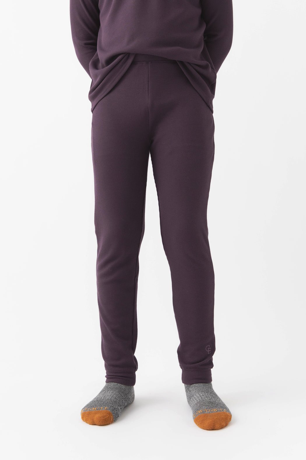 A child stands against a white background wearing Cozy Earth’s Children’s Bamboo Waffle Knit Base Layer Pant in Plum, paired with a matching top and gray socks with orange toes. Their hands are in their pockets, upper body partially out of frame.|Color:Plum
