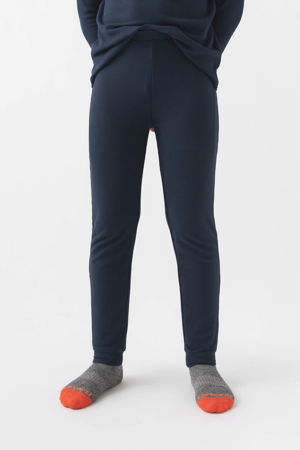 A child wears Cozy Earth Children's Bamboo Waffle Knit Base Layer Pant in navy, paired with gray socks with orange toes, standing against a plain white background. The upper body is mostly out of the frame.|Color:Navy