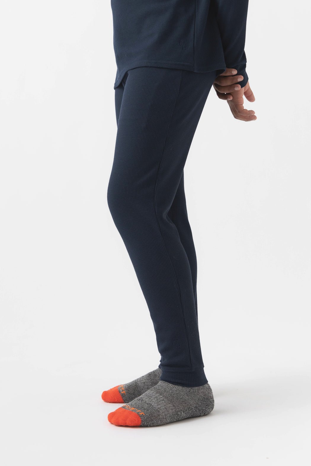 A child’s lower body is shown on a white background, wearing Cozy Earth Children’s Bamboo Waffle Knit Base Layer Pants in navy, a matching long-sleeve top, and gray socks with bright orange toes and heels.|Color:Navy