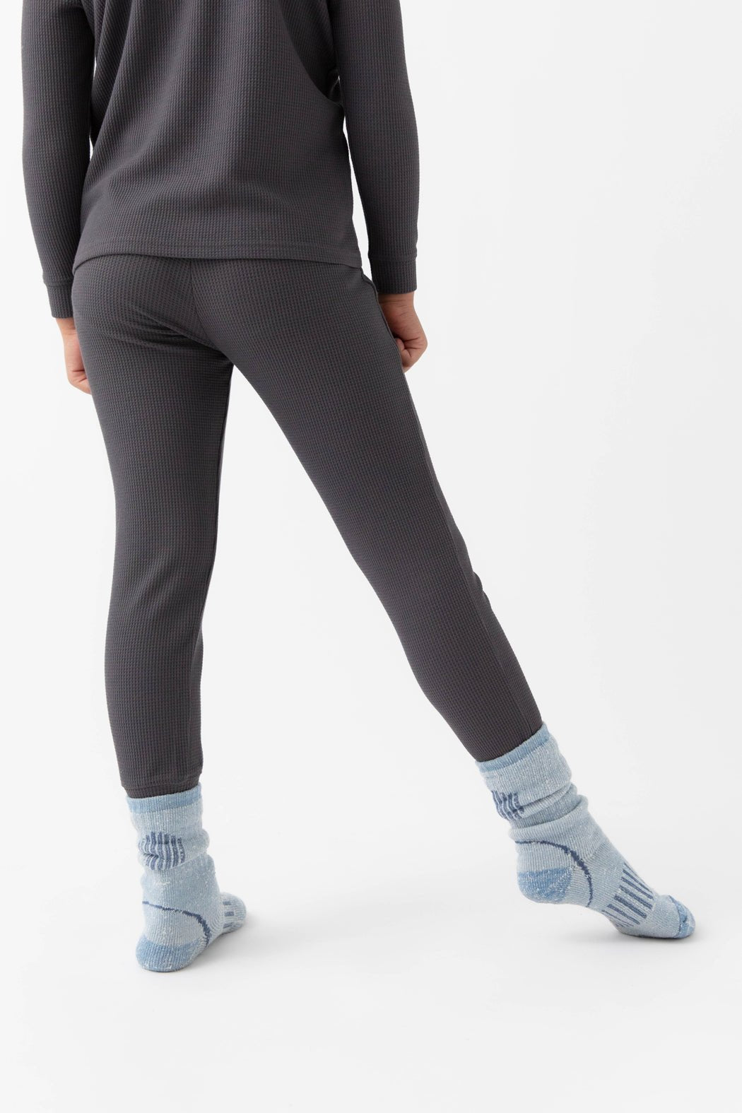 A child stands with their back turned, wearing Cozy Earth's Children's Bamboo Waffle Knit Base Layer Pant in dark ribbed fabric, paired with a matching top and light blue slouchy socks, set against a plain white background.|Color:Iron