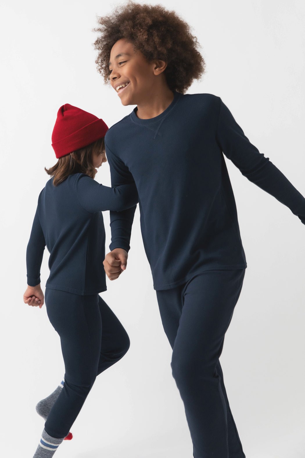 Two children wearing matching Cozy Earth Children's Bamboo Waffle Knit Base Layer Long Sleeves, one with a red beanie, happily holding hands and walking together against a plain white background.|Color:Navy