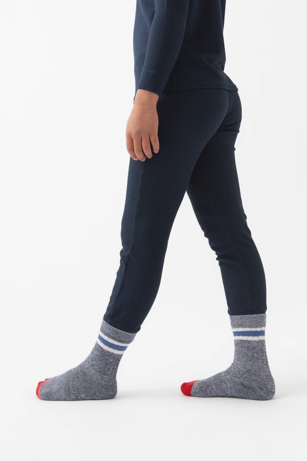 A child models the Children's Bamboo Waffle Knit Base Layer Pant by Cozy Earth, standing sideways in navy pajamas and gray socks with white stripes and red toes against a plain white background.|Color:Navy