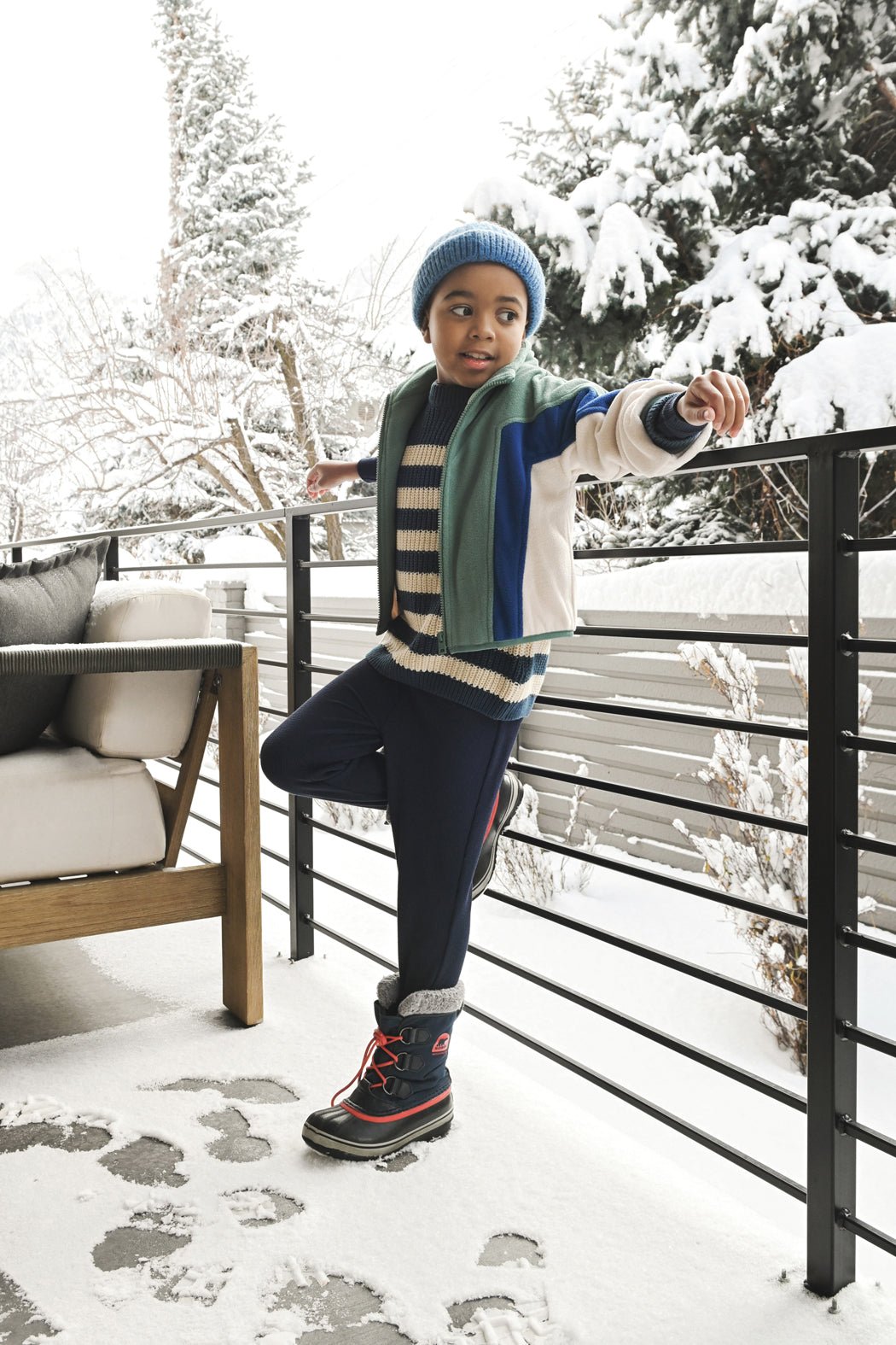 A child smiles and looks to the side on a snowy patio, dressed in Cozy Earth's Children's Bamboo Waffle Knit Base Layer Pant, winter layers, and a blue beanie. Snow-covered trees and furniture appear in the background.|Color:Navy