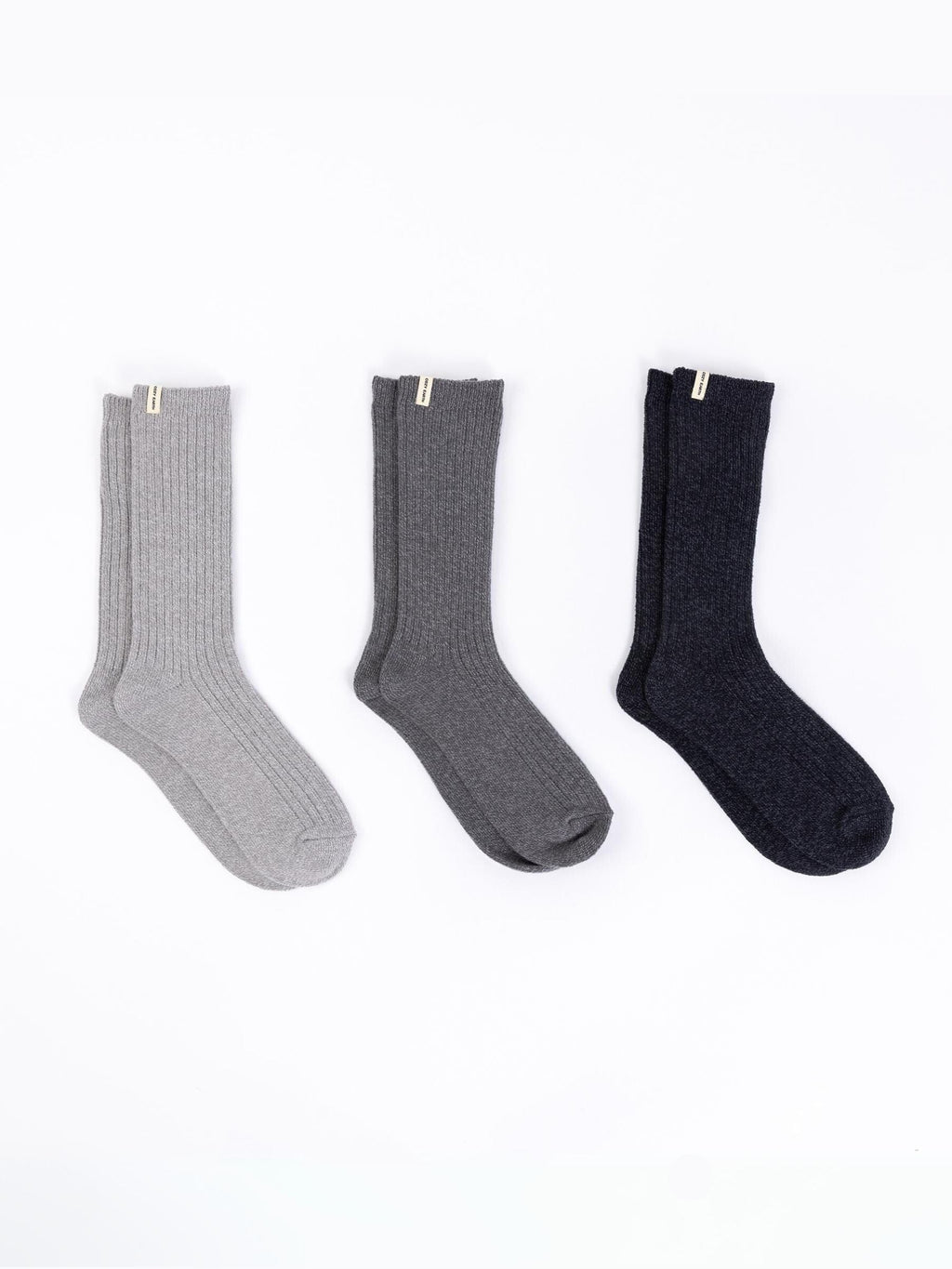 The Plush Lounge Calf Sock 3-Pack from Cozy Earth features three pairs of ribbed socks displayed against a white backdrop, showcasing a palette of light gray, dark gray, and black. Each pair is adorned with a small tag at the top. |Color:Charcoal/Coal/Stone