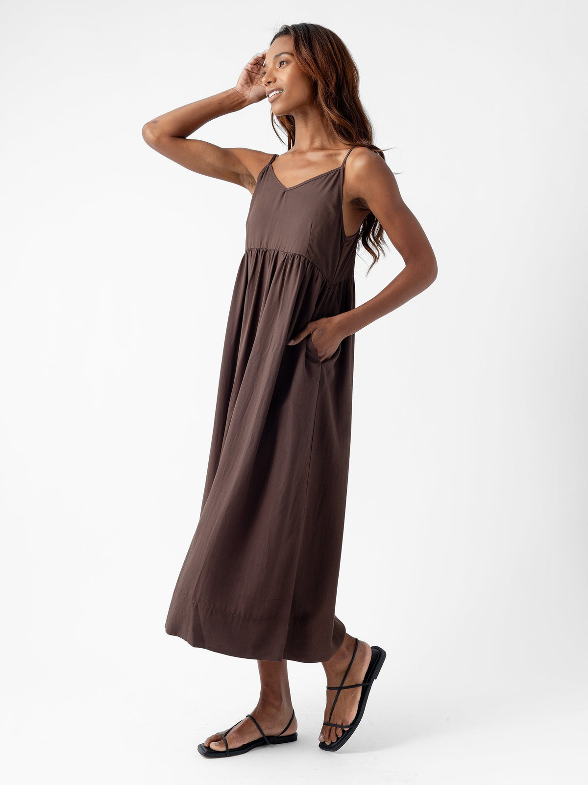A person stands against a white background wearing the Women's Sunset Gathered Dress by Cozy Earth. The sleeveless, brown maxi dress features convenient pockets. They are smiling and looking off to the side with one hand raised to their head and the other resting on a pocket. They are wearing black sandals, and their long hair flows down. |Color:Cacao