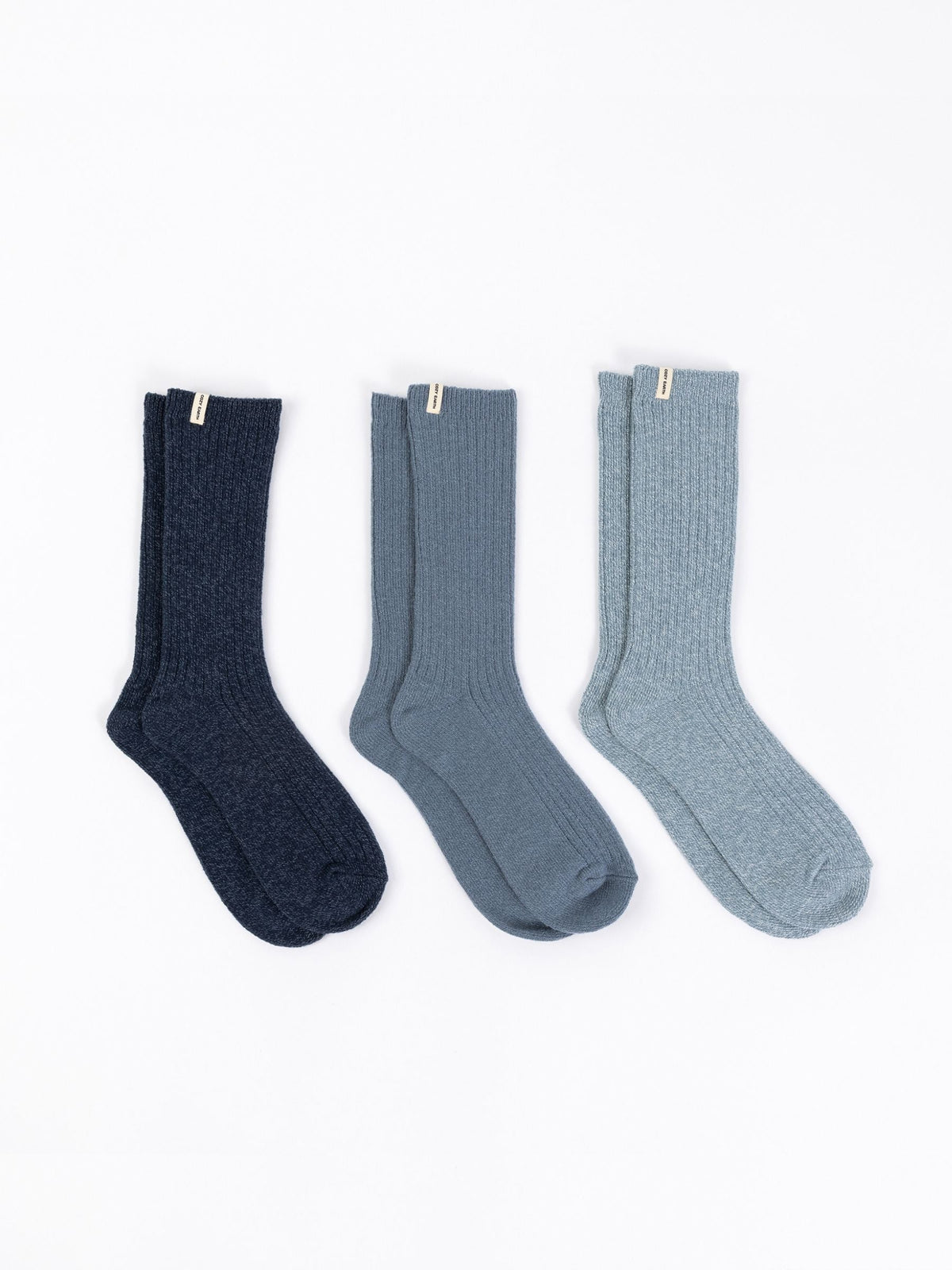 A set of The Plush Lounge Calf Sock 3-Pack by Cozy Earth, featuring three pairs in various blue tones, is stylishly displayed on a white background, each with a small tag near the cuff. |Color:Eclipse/Deep Blue/Smokey Blue