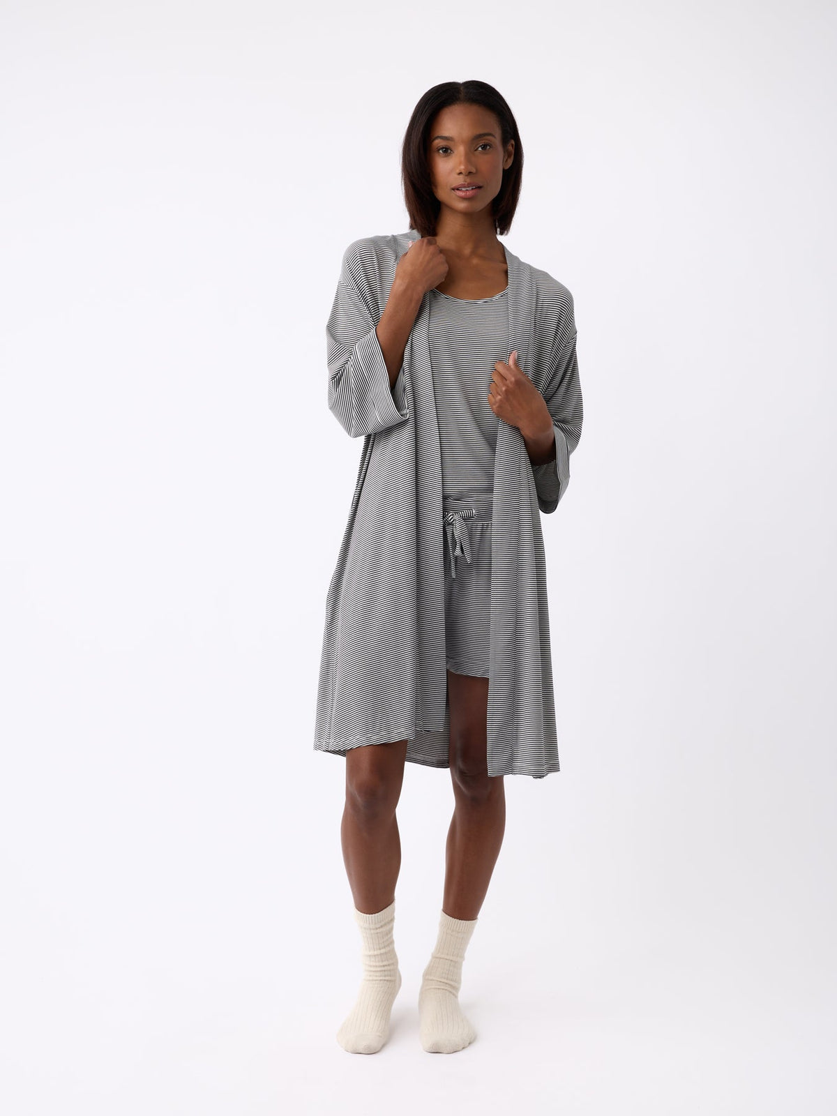 Against a plain backdrop, a person wears a Cozy Earth Women's Bamboo Stretch-Knit Kimono Robe in light gray over a matching top and shorts. They pair it with cream socks and have shoulder-length hair. |Color:Eclipse Mini Stripe