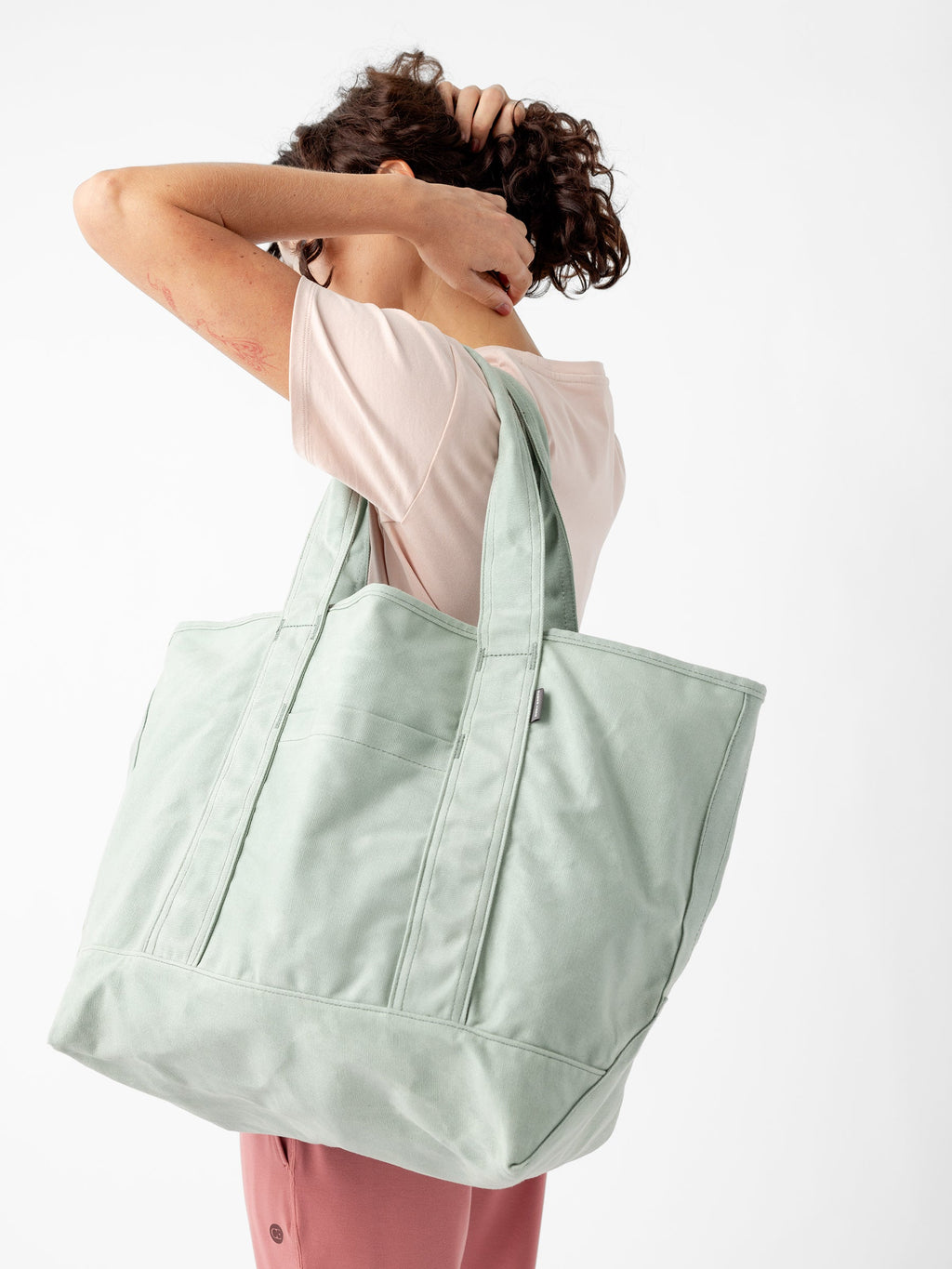 A person with short, curly hair is scratching their head while holding a large, light green Waxed Canvas Tote from Cozy Earth over their shoulder. They are wearing a light pink T-shirt and pink pants, standing against a plain white background. |Color:Palm