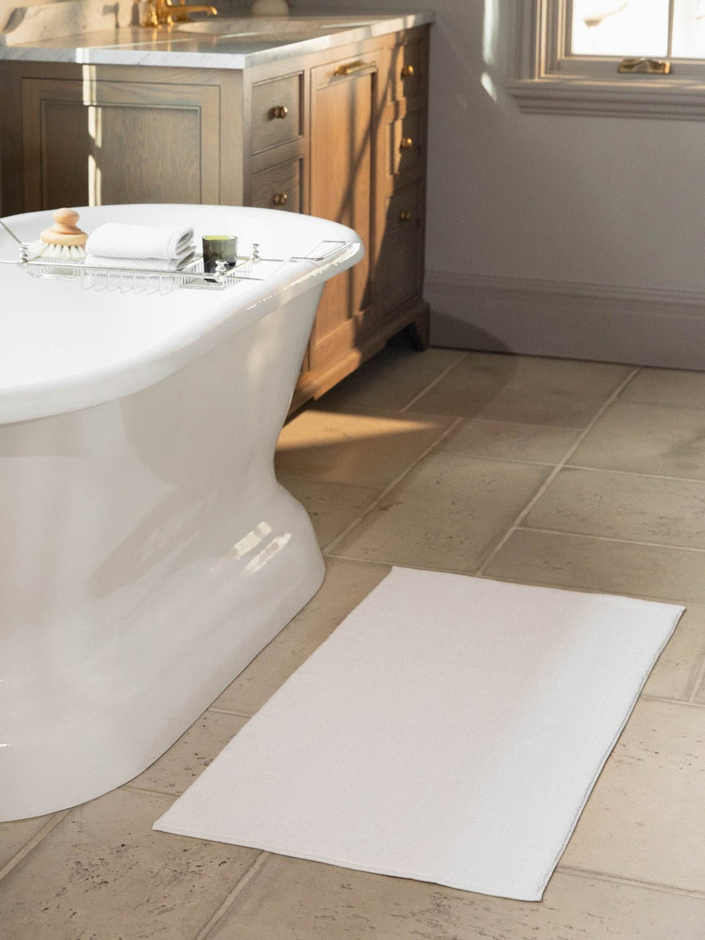 The Cozy Earth Loop Terry Bath Mat in white rests on a beige tiled floor beside a freestanding tub, with a wooden vanity with drawers and a window visible in the background.|Color:White