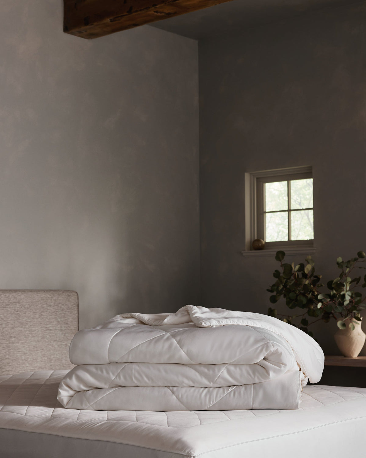 A Cozy Earth Bamboo Insert Bundle is neatly folded on a bed in a softly lit, minimalist bedroom with grey walls, a small window, and a ceramic vase with greenery on a wooden nightstand.|Filling:Extra