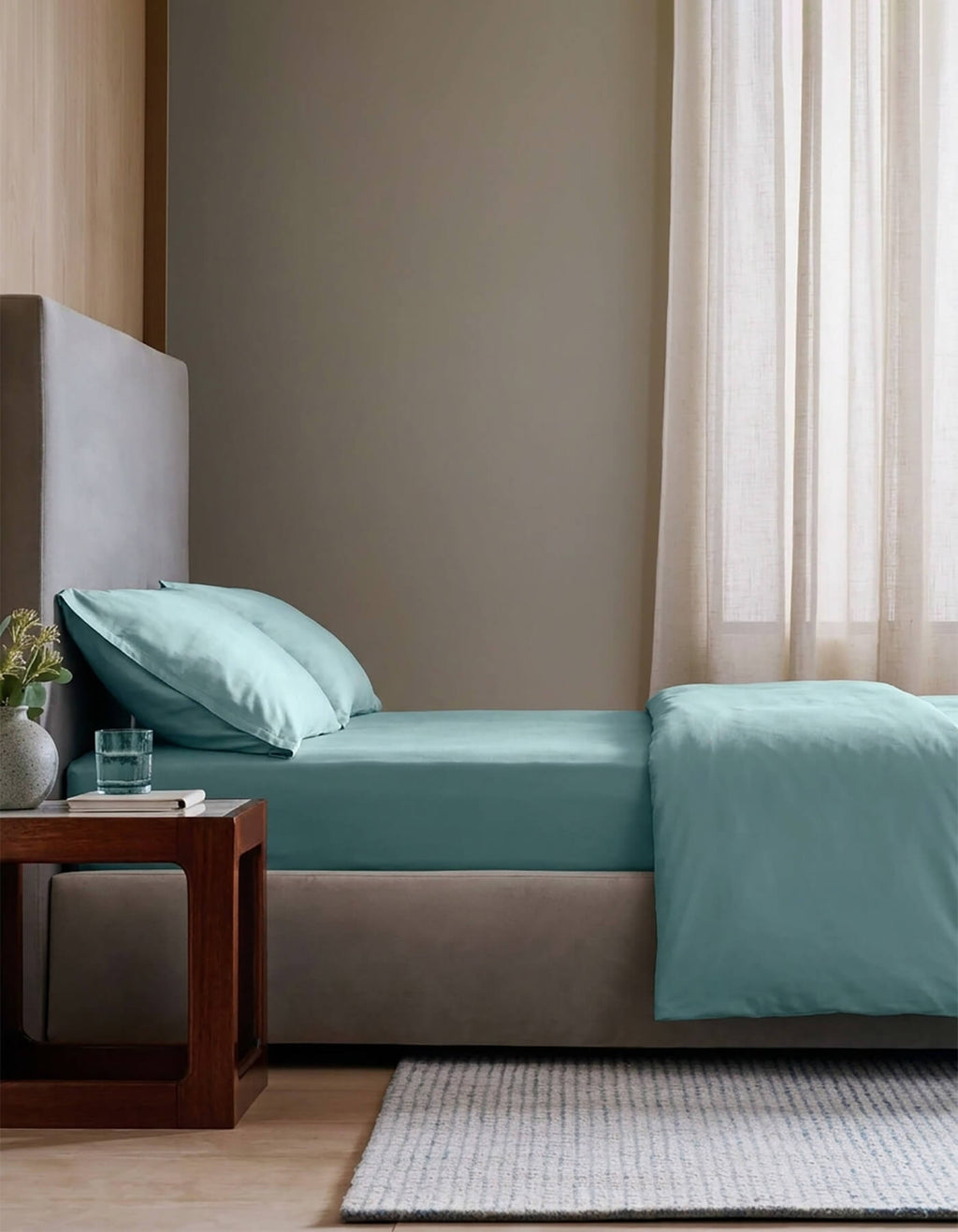 A modern bedroom with a neatly made bed featuring the Cozy Earth Bamboo Bedding Essential Bundle, light blue sheets and pillows, a wooden nightstand with a glass of water and plant, beige walls, and sheer curtains letting in natural light.|Color:River
