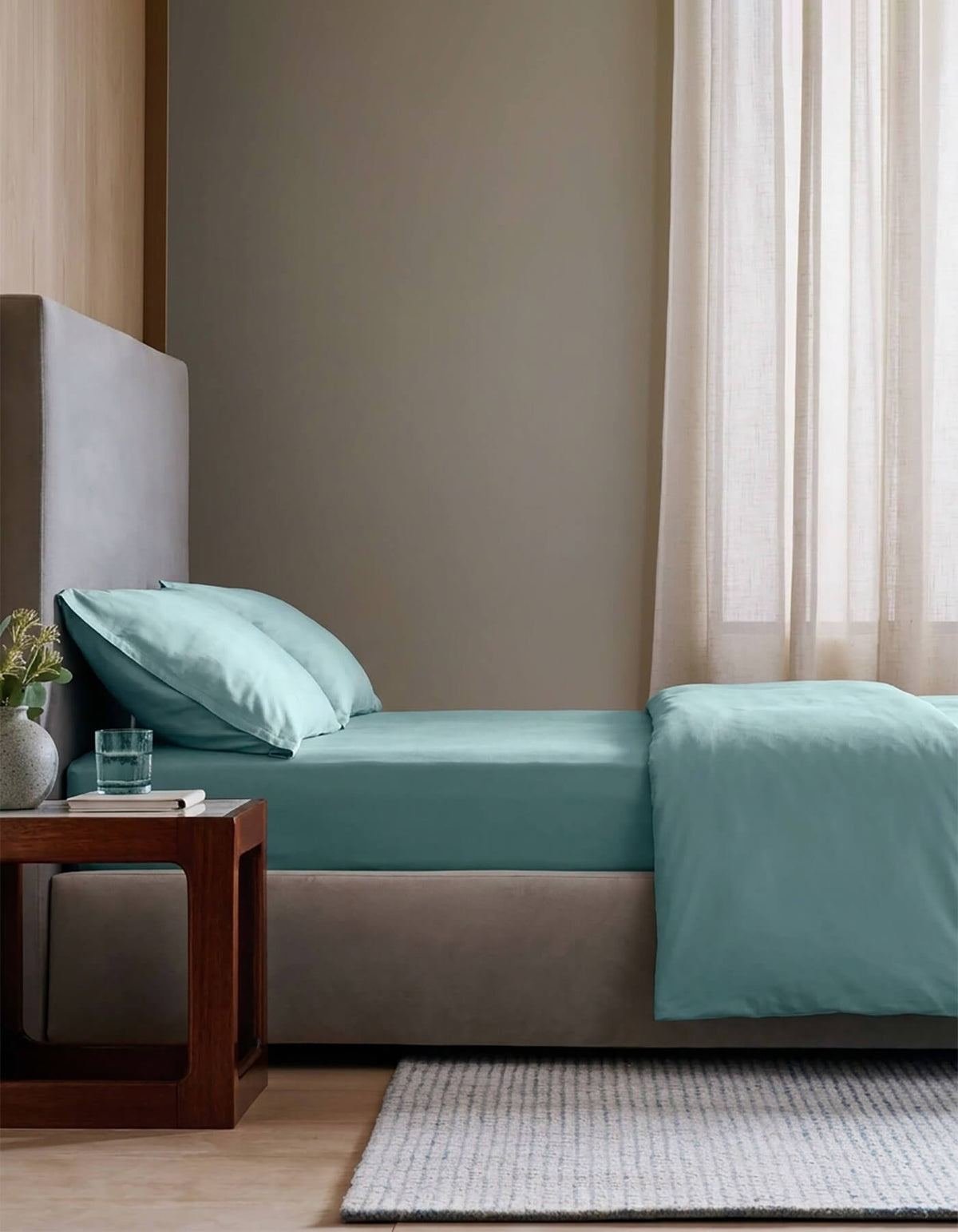 A modern bedroom with a neatly made bed featuring the Cozy Earth Bamboo Bedding Essential Bundle, light blue sheets and pillows, a wooden nightstand with a glass of water and plant, beige walls, and sheer curtains letting in natural light.|Color:River