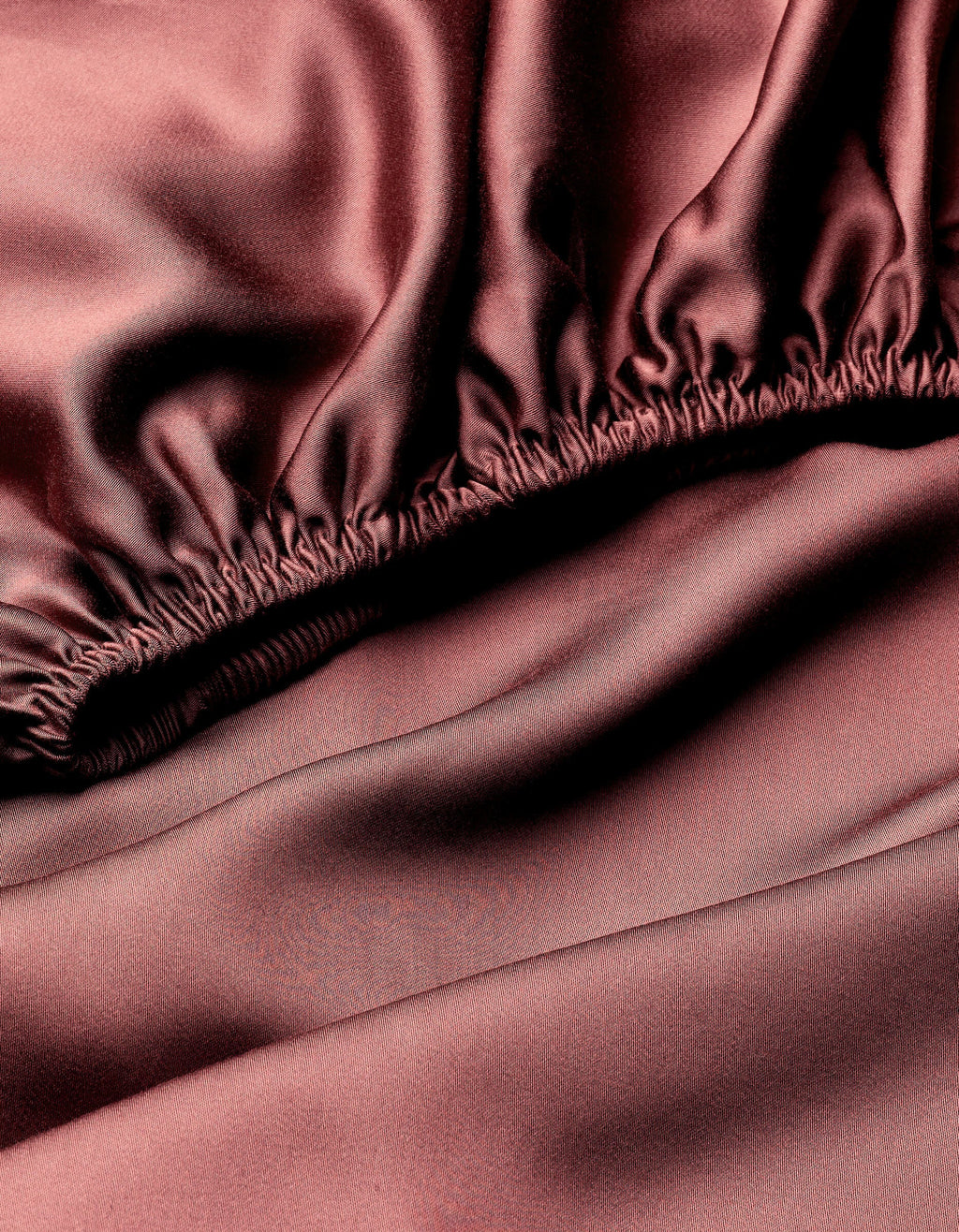 Close-up of Cozy Earth's Bamboo Fitted Sheet in smooth, silky dark pink fabric with gathered elastic edge and a shiny, luxurious finish.|Color:Mulberry
