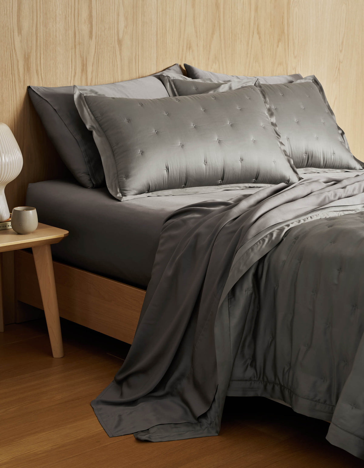 A neatly made bed features the Cozy Earth Bamboo Quilt Bundle—gray, silky bedding with embroidered pillows—beside a wooden nightstand holding a white lamp and mug, set against a light wood-paneled wall.|Color:Charcoal