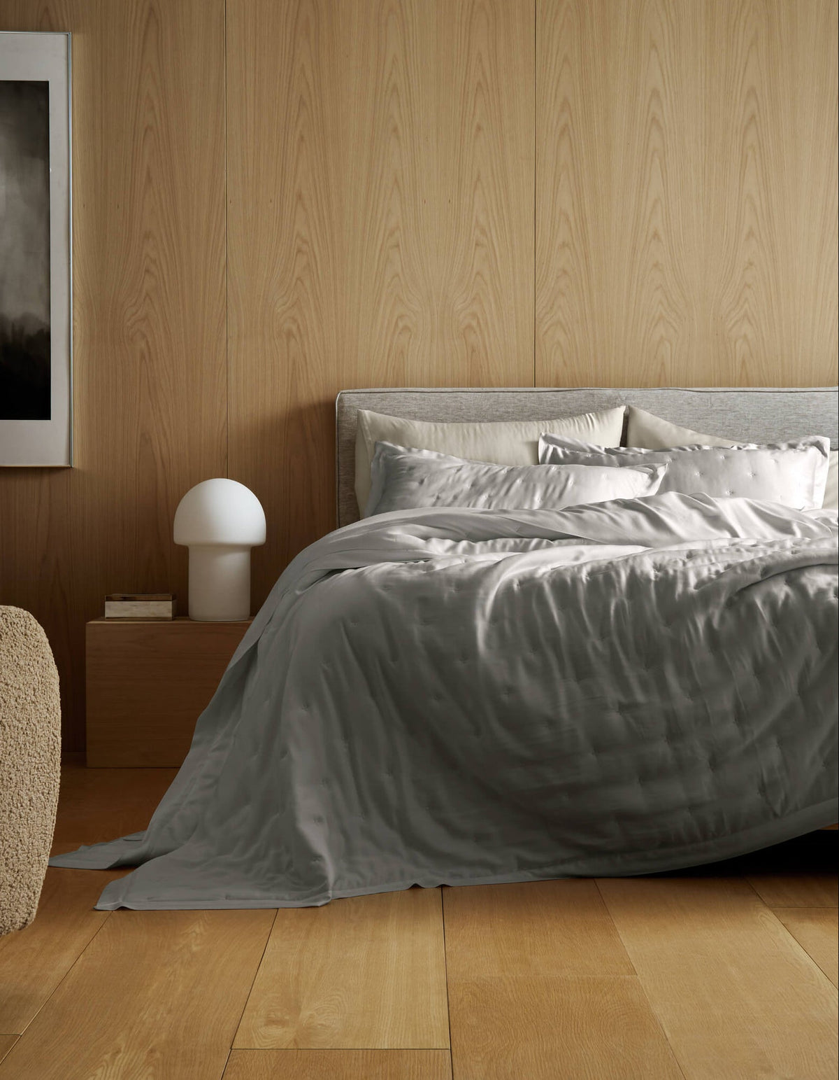 A modern bedroom featuring a light wood floor and wall, a neatly made bed with the Cozy Earth Bamboo Quilt Bundle in silver-gray, a wooden nightstand with a white lamp, a beige textured chair, and abstract wall art.|Color:Light Grey
