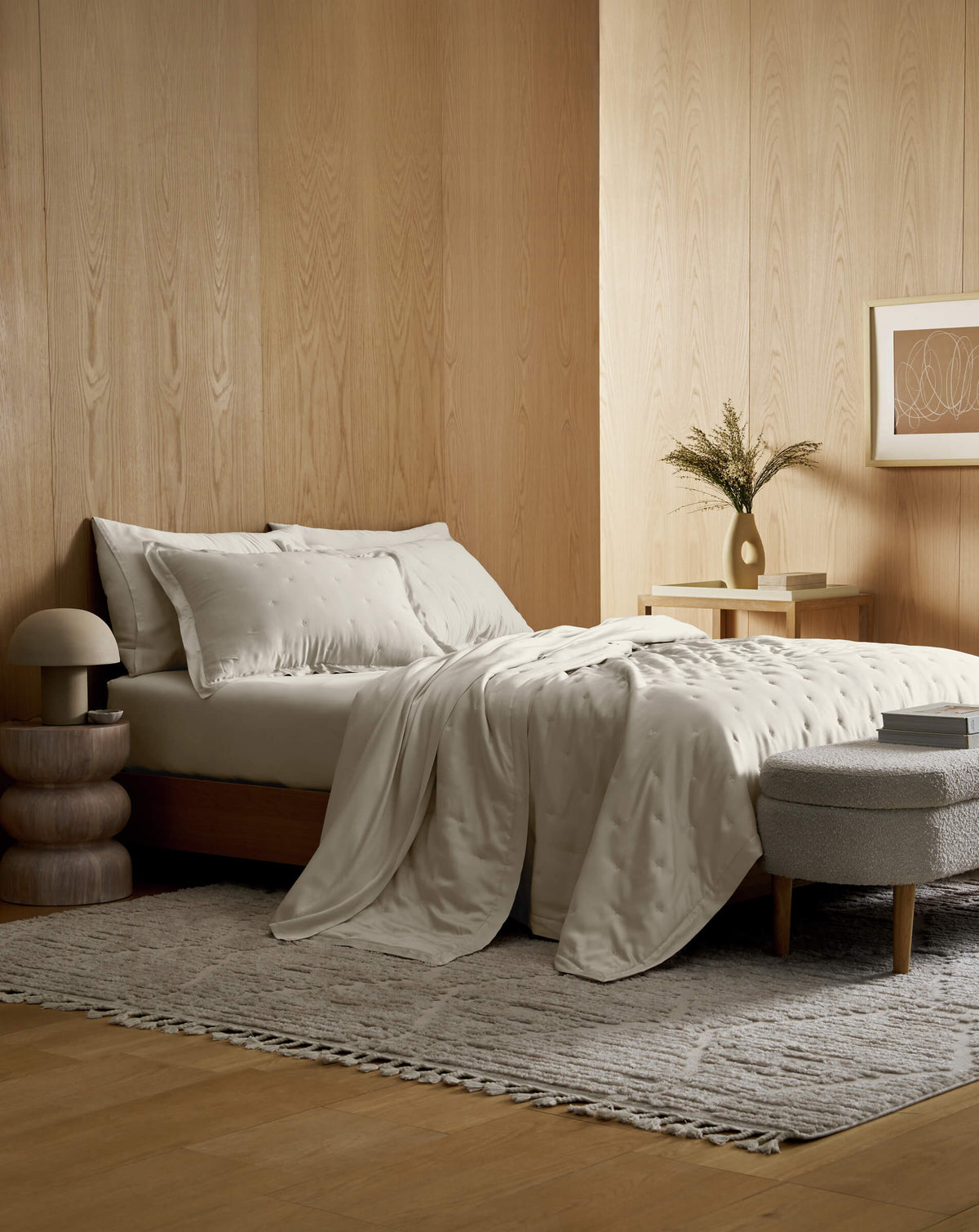 A minimalist bedroom with light wood walls, a neatly made bed featuring the Cozy Earth Bamboo Quilt Bundle, a small nightstand with a vase and dried flowers, a textured rug, round stool, and framed art on the wall.|Color:Oat