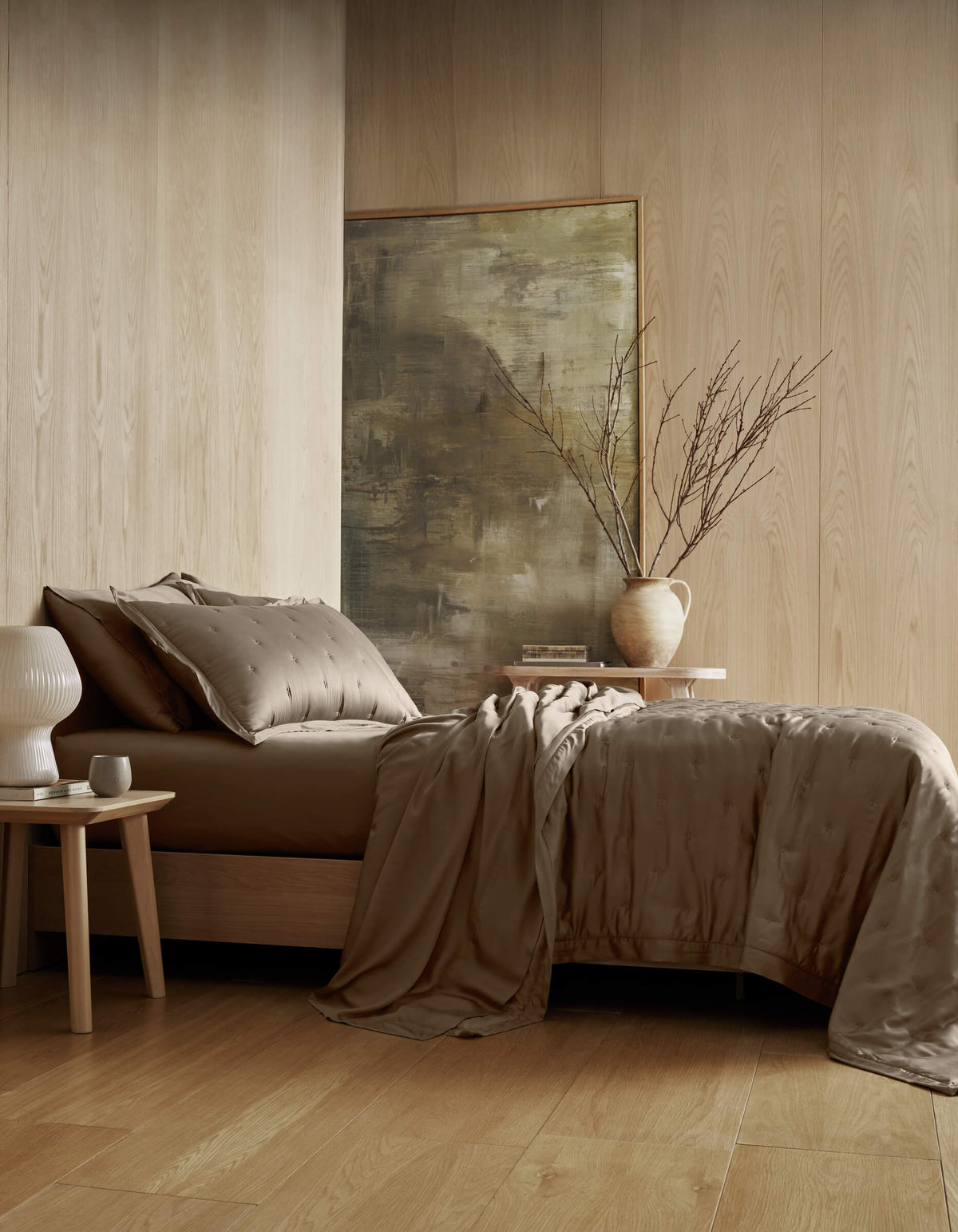 A modern bedroom features the Cozy Earth Bamboo Quilt Bundle with taupe bedding and matching pillows. Decorative touches include a beige vase with branches, a white lamp, and a small side table against wooden paneling and an abstract painting.|Color:Walnut