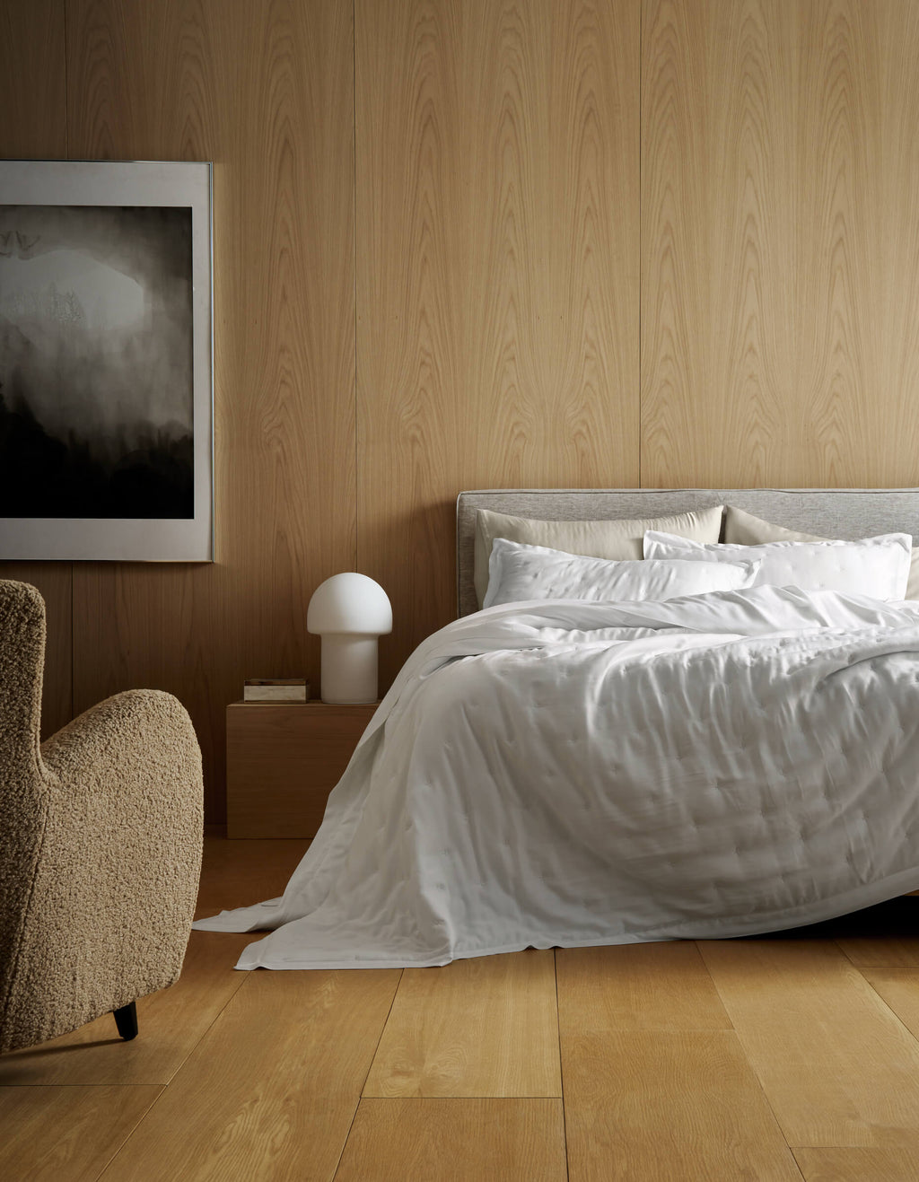 A modern bedroom features light wood panels, a bed made with Cozy Earth’s Bamboo Quilt, a beige armchair, a wooden nightstand with a white lamp, and abstract black-and-white artwork on the wall.|Color:White