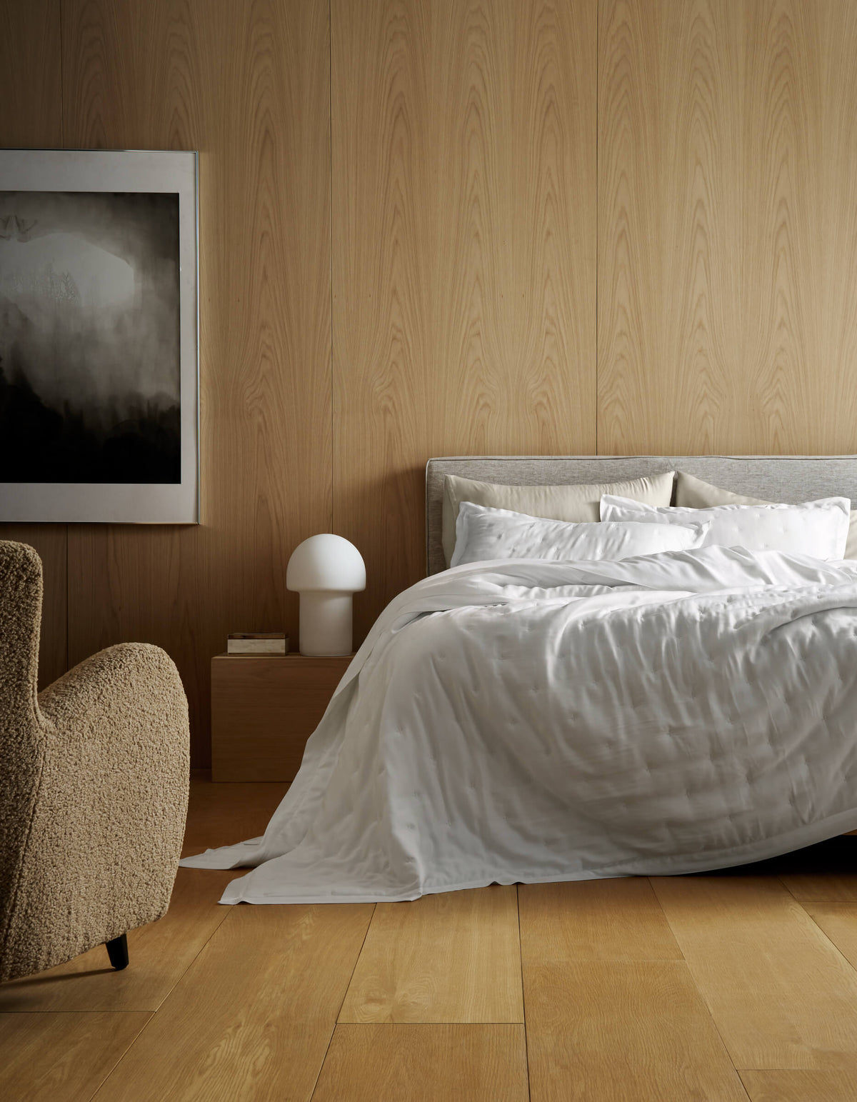 A modern bedroom features light wood panels, a bed made with Cozy Earth’s Bamboo Quilt, a beige armchair, a wooden nightstand with a white lamp, and abstract black-and-white artwork on the wall.|Color:White