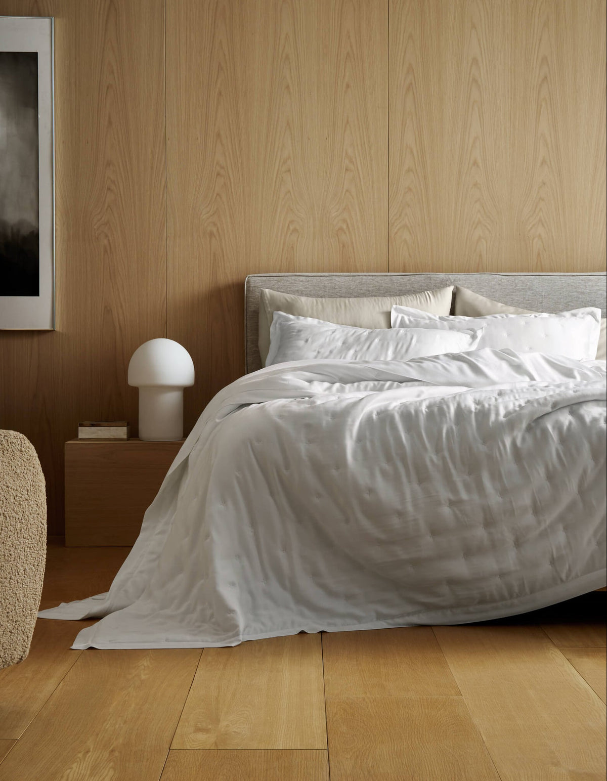 A modern bedroom features light wood-paneled walls, a large bed with Cozy Earth’s Bamboo Quilt Bundle, a beige chair, wooden nightstand with a white mushroom lamp, books, and an abstract painting on the wall.|Color:White