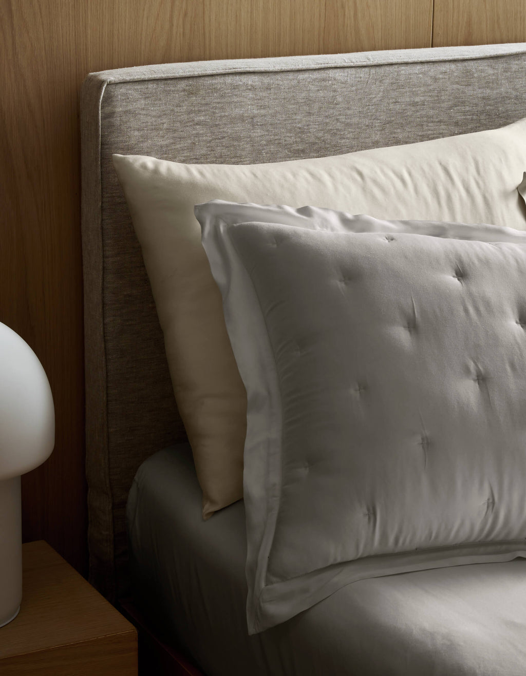 A neatly made bed with Cozy Earth Bamboo Quilted Shams, a padded fabric headboard, and a gray sheet. A modern white lamp is on a wooden nightstand beside the bed.|Color:Light Grey