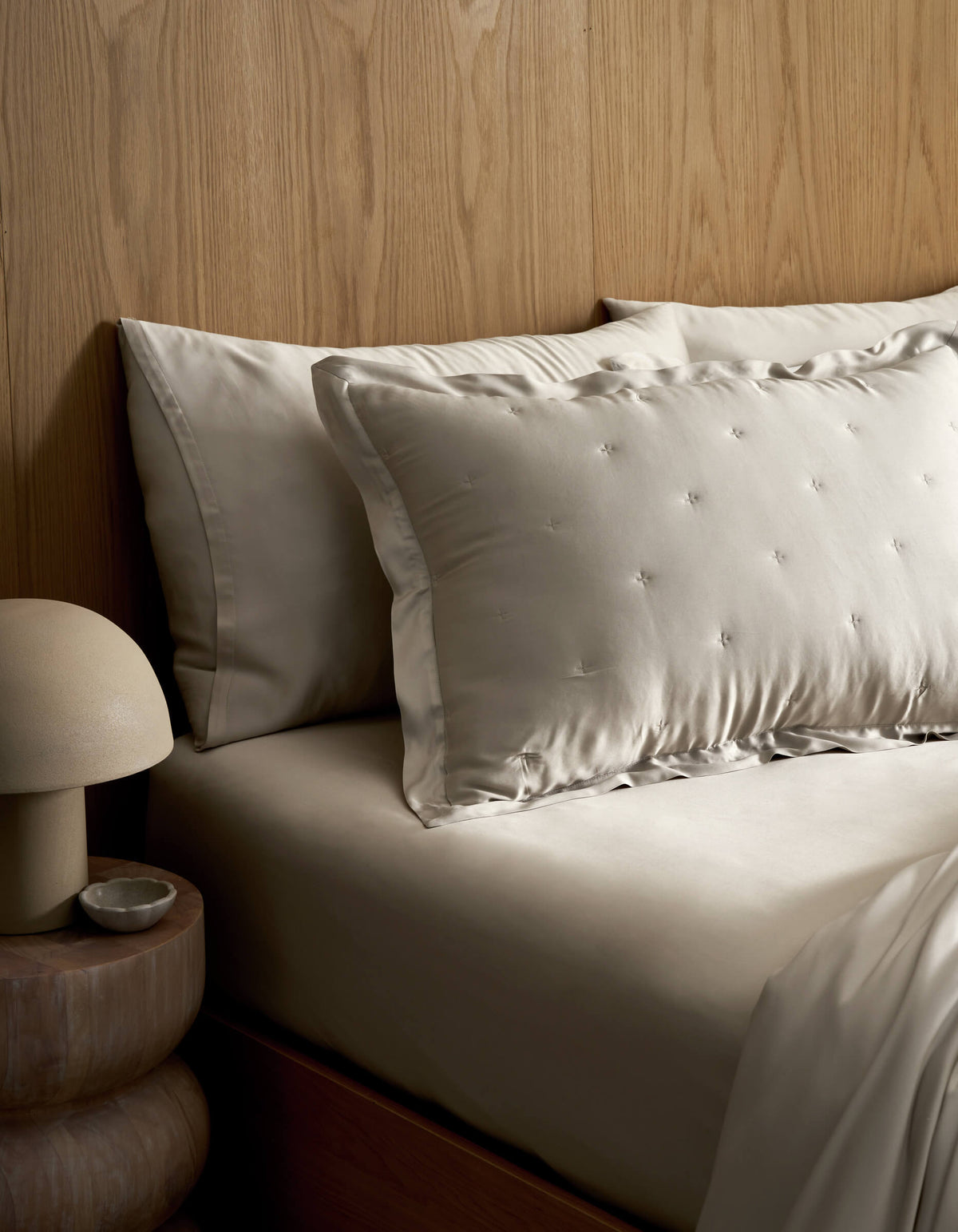 A neatly made bed features Cozy Earth's Bamboo Quilted Shams in light tones, set against a wooden headboard. A round bedside table holds a dome-shaped lamp and a small dish.|Color:Oat