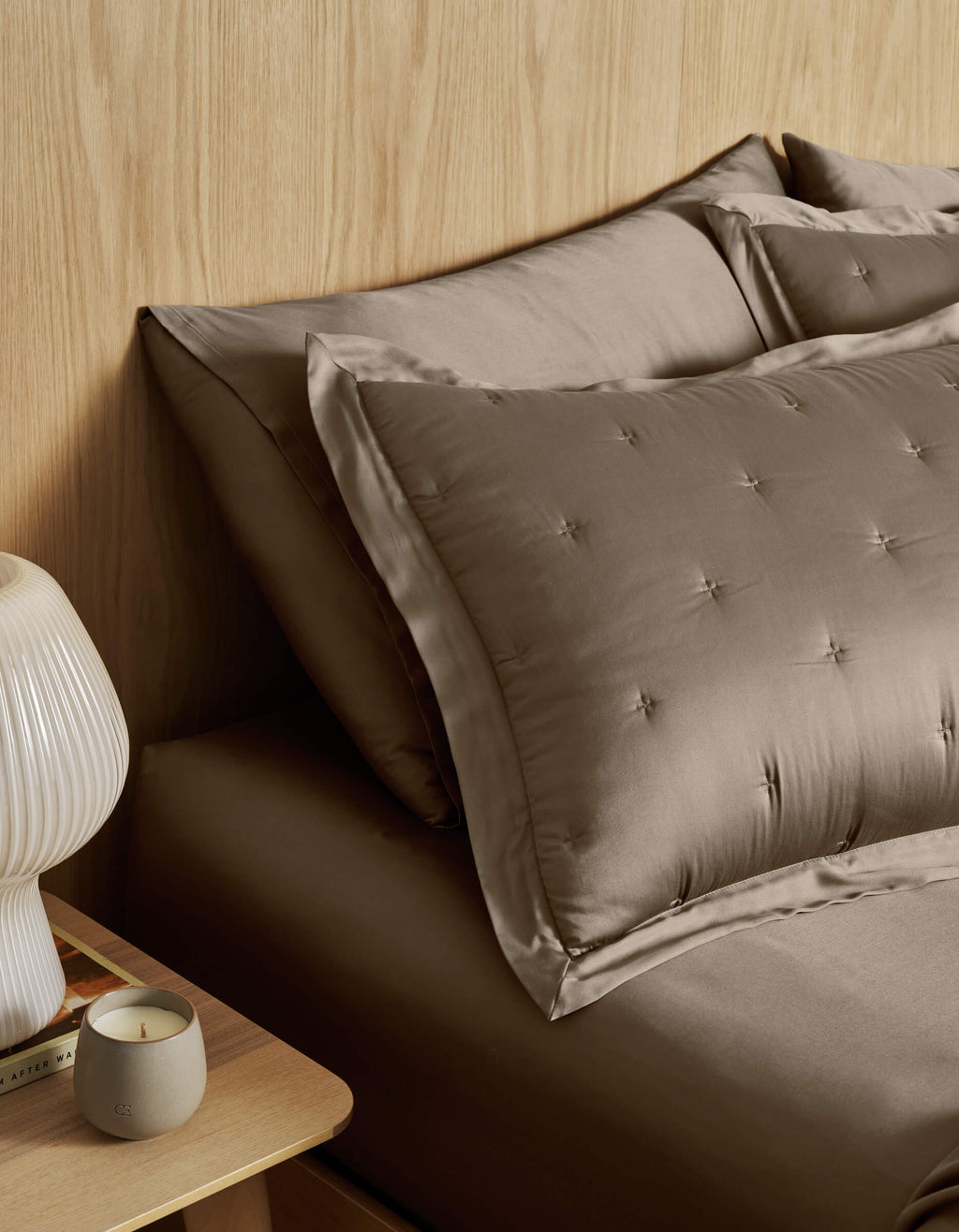 A neatly made bed with Cozy Earth Bamboo Quilted Shams and taupe satin bedding sits by a wooden nightstand topped with a ribbed white lamp, a lit candle, and a book against a light wood-paneled wall.|Color:Walnut