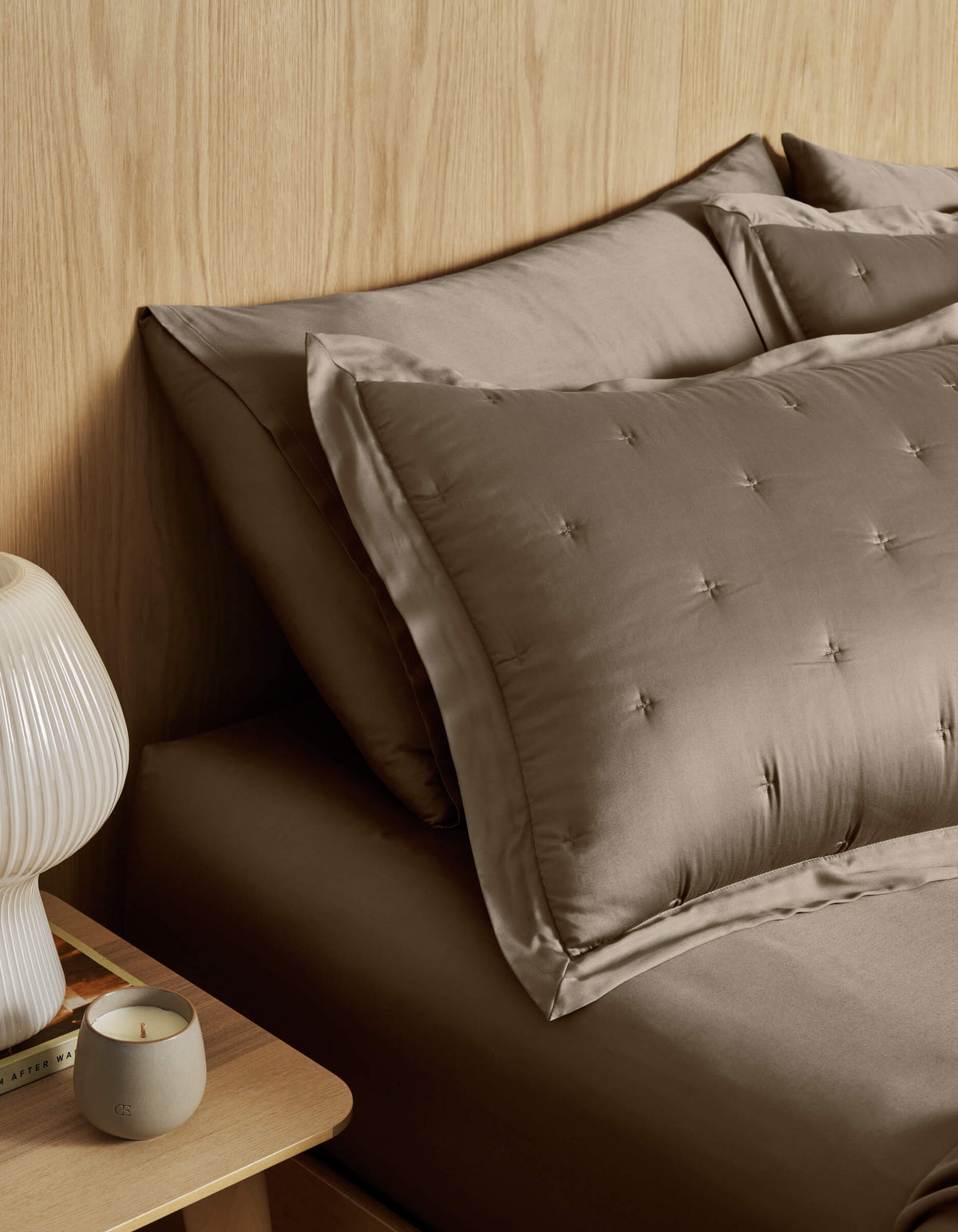A neatly made bed with Cozy Earth Bamboo Quilted Shams and taupe satin bedding sits by a wooden nightstand topped with a ribbed white lamp, a lit candle, and a book against a light wood-paneled wall.|Color:Walnut