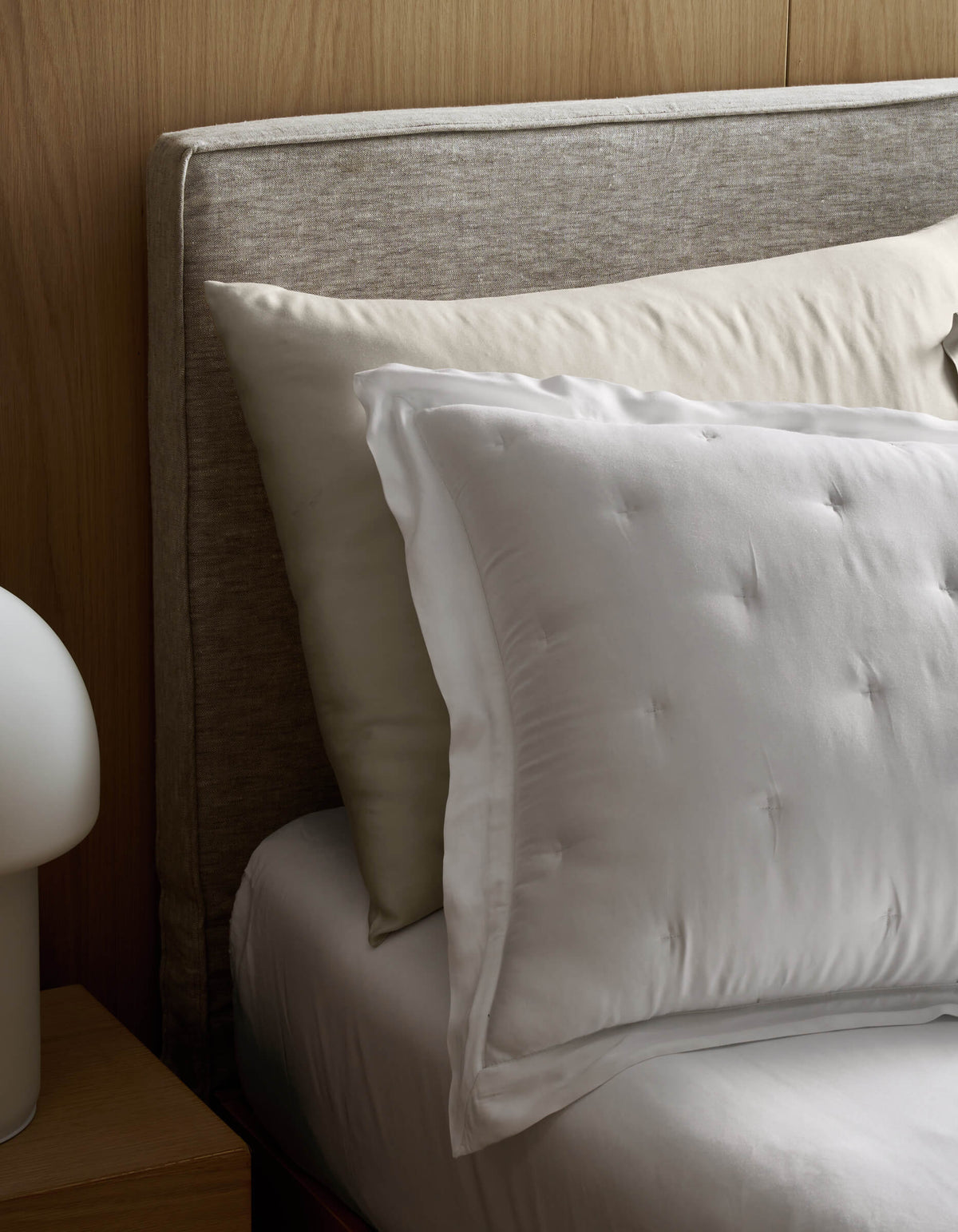 A close-up of a bed featuring Cozy Earth Bamboo Quilted Shams, paired with cream and white pillows, white sheets, a light gray upholstered headboard, and part of a round white bedside lamp on a wooden nightstand.|Color:White