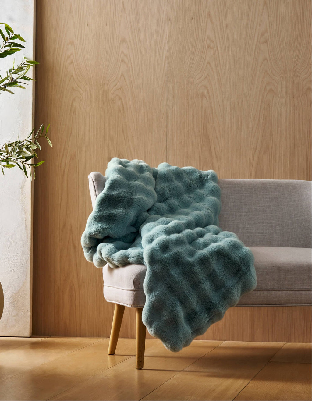 A light gray chair with wooden legs displays the Bubble Cuddle Blanket by Cozy Earth in textured teal, set in a room with light wood-paneled walls, a potted plant, and a white canvas. Natural light brightens the space.|Color:Tide