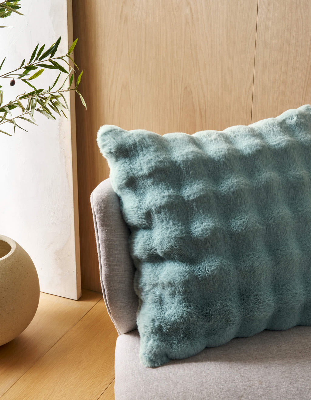 A close-up of the Cozy Earth Bubble Cuddle Pillow in light blue, with a fluffy texture, rests on a light grey sofa beside a round beige planter with green leafy branches, set against a light wood-paneled wall.|Color:Tide |Size:Euro