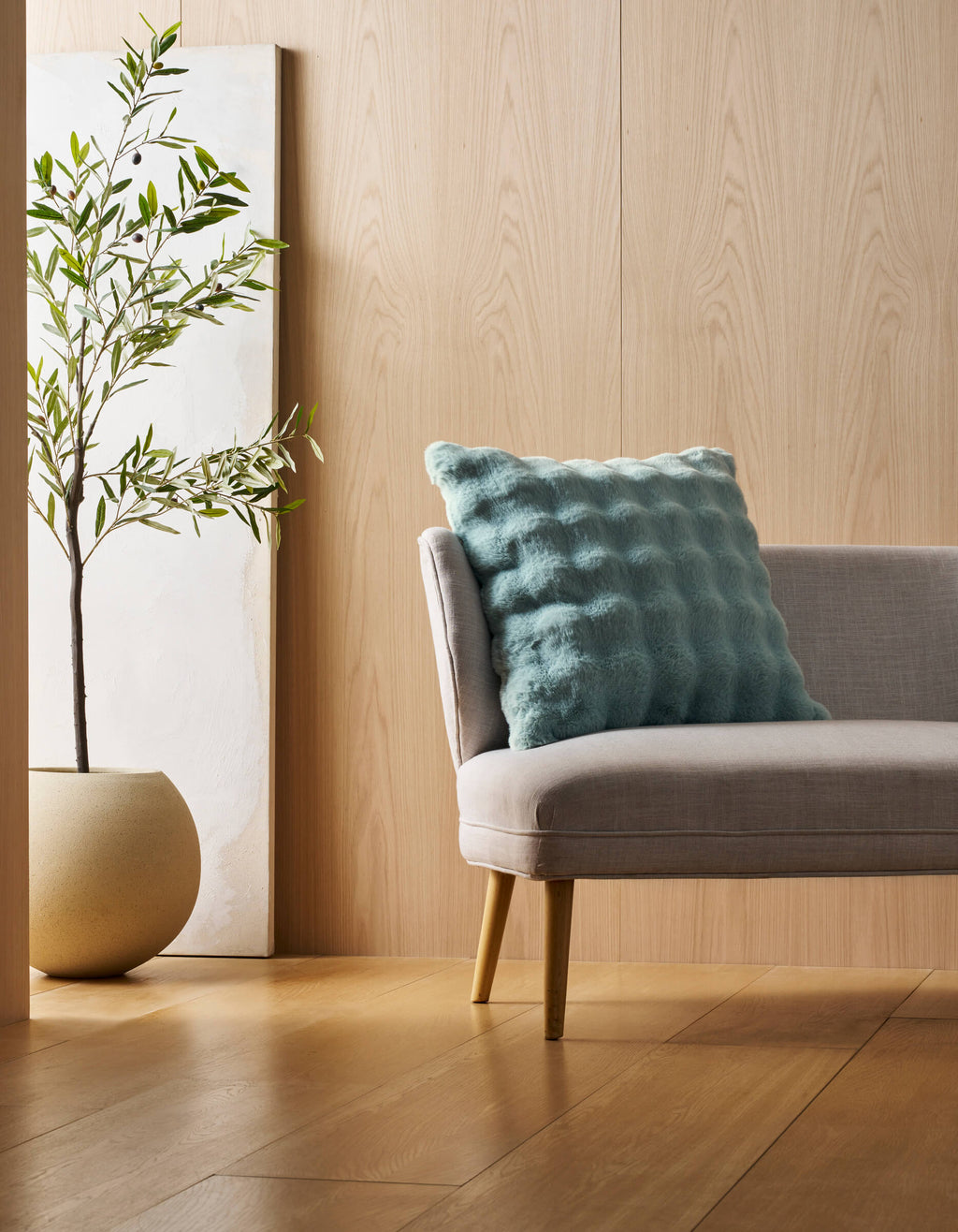 A Bubble Cuddle Blanket by Cozy Earth is draped over a light gray chair with a blue pillow, set on a wooden floor near a potted plant and white panel against a light wood-paneled wall in a minimalist room.|Color:Tide |Size:Euro