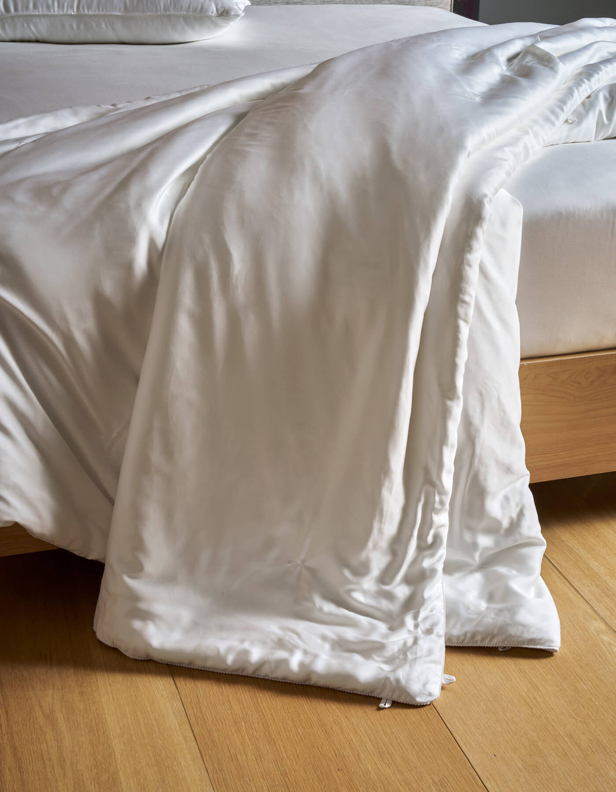 A Cozy Earth Silk Comforter drapes over the edge of a light wooden bed frame onto a wooden floor, with matching white bedding visible on the bed.|Filling:Extra