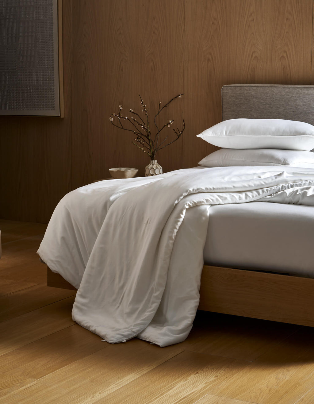A neatly made bed with a gray headboard and white bedding features the Cozy Earth Silk Comforter draped over the edge. A vase with branches sits on the bedside table, all set on wooden floors with a wood-paneled wall in the background.|Filling:Extra