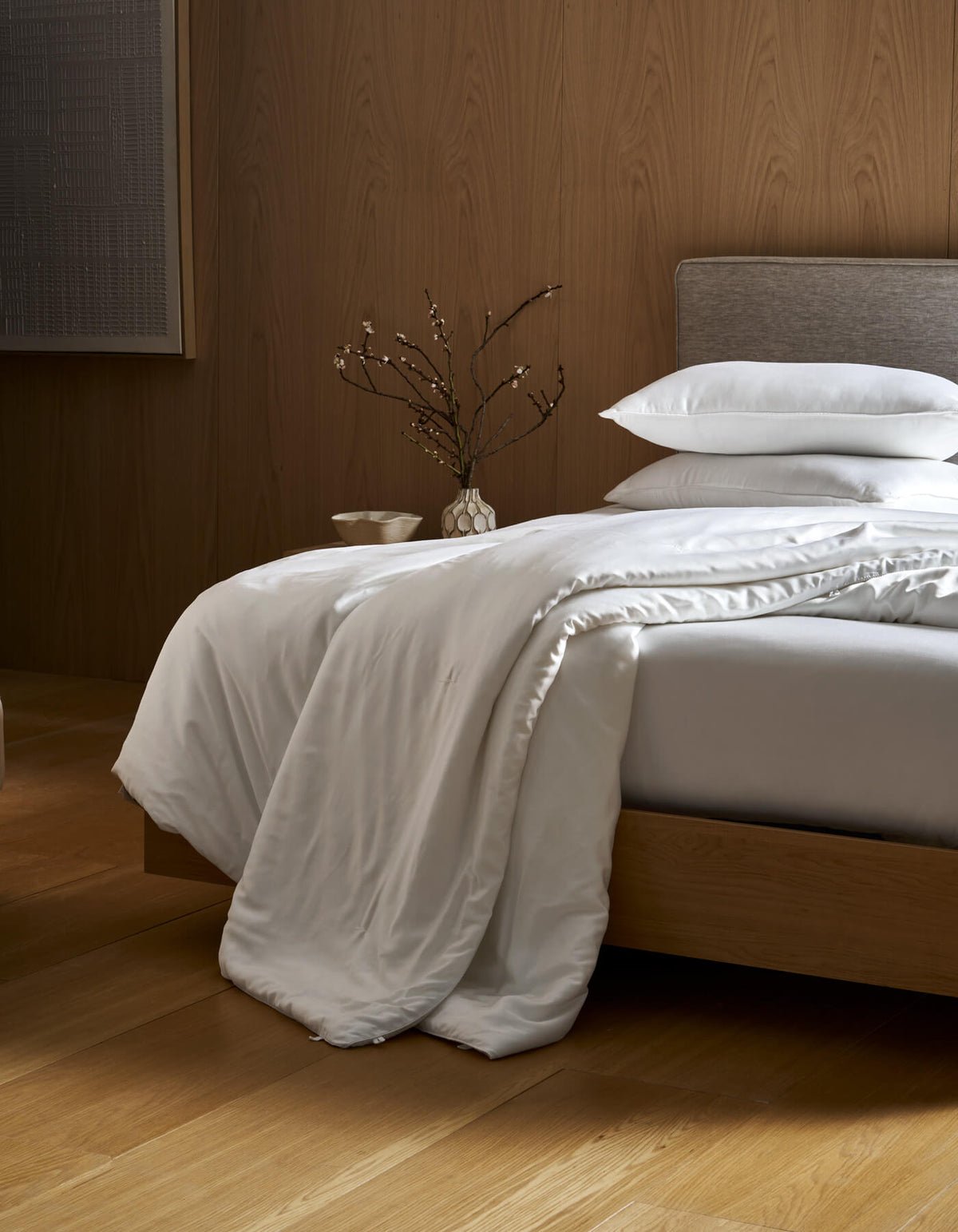 A neatly made bed with a gray headboard and white bedding features the Cozy Earth Silk Comforter draped over the edge. A vase with branches sits on the bedside table, all set on wooden floors with a wood-paneled wall in the background.|Filling:Extra