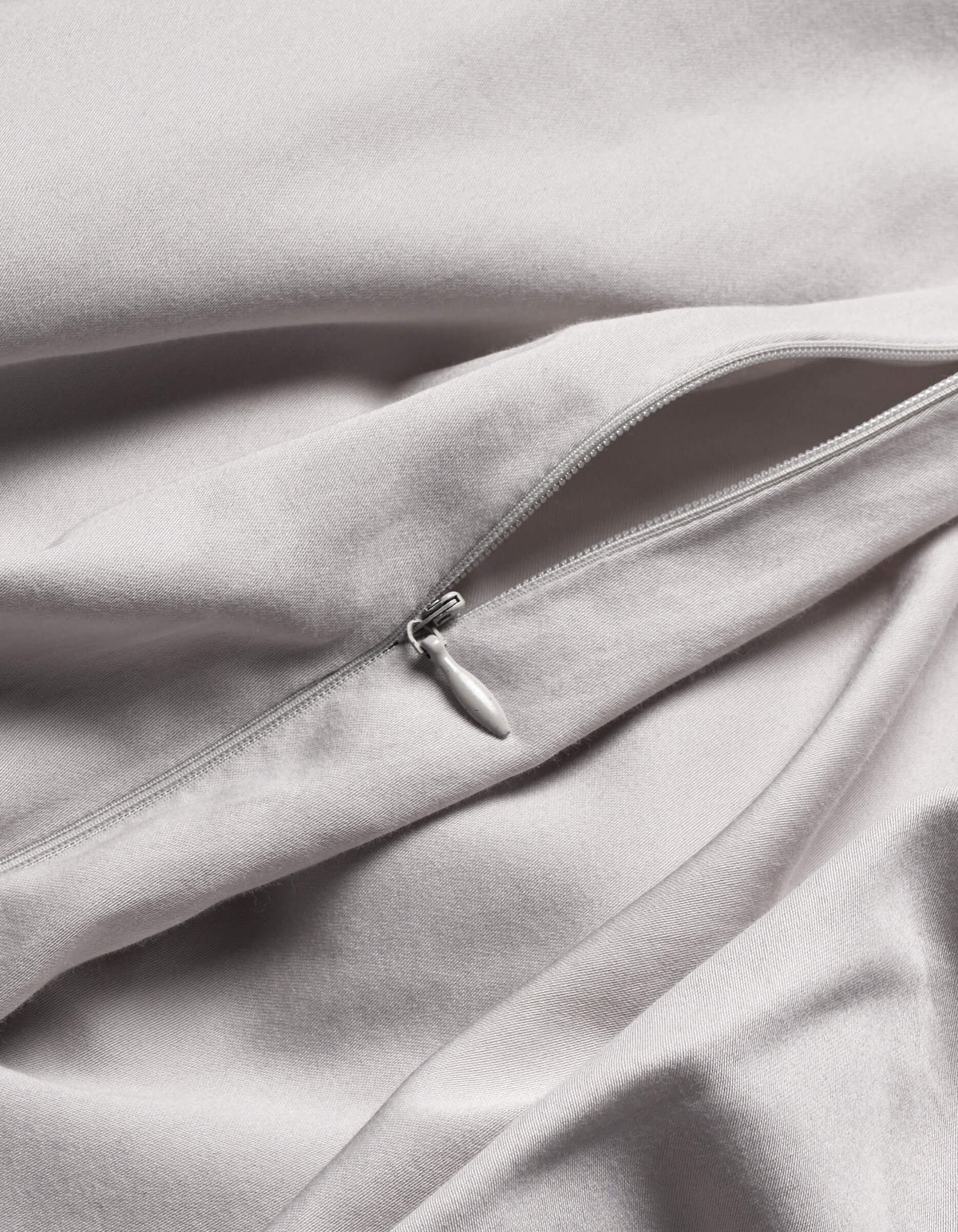 A close-up of Cozy Earth's Soft-Wash Cotton Duvet Cover in light grey shows a silver zipper partially unzipped on its smooth, gently folded fabric.|Color:Dew