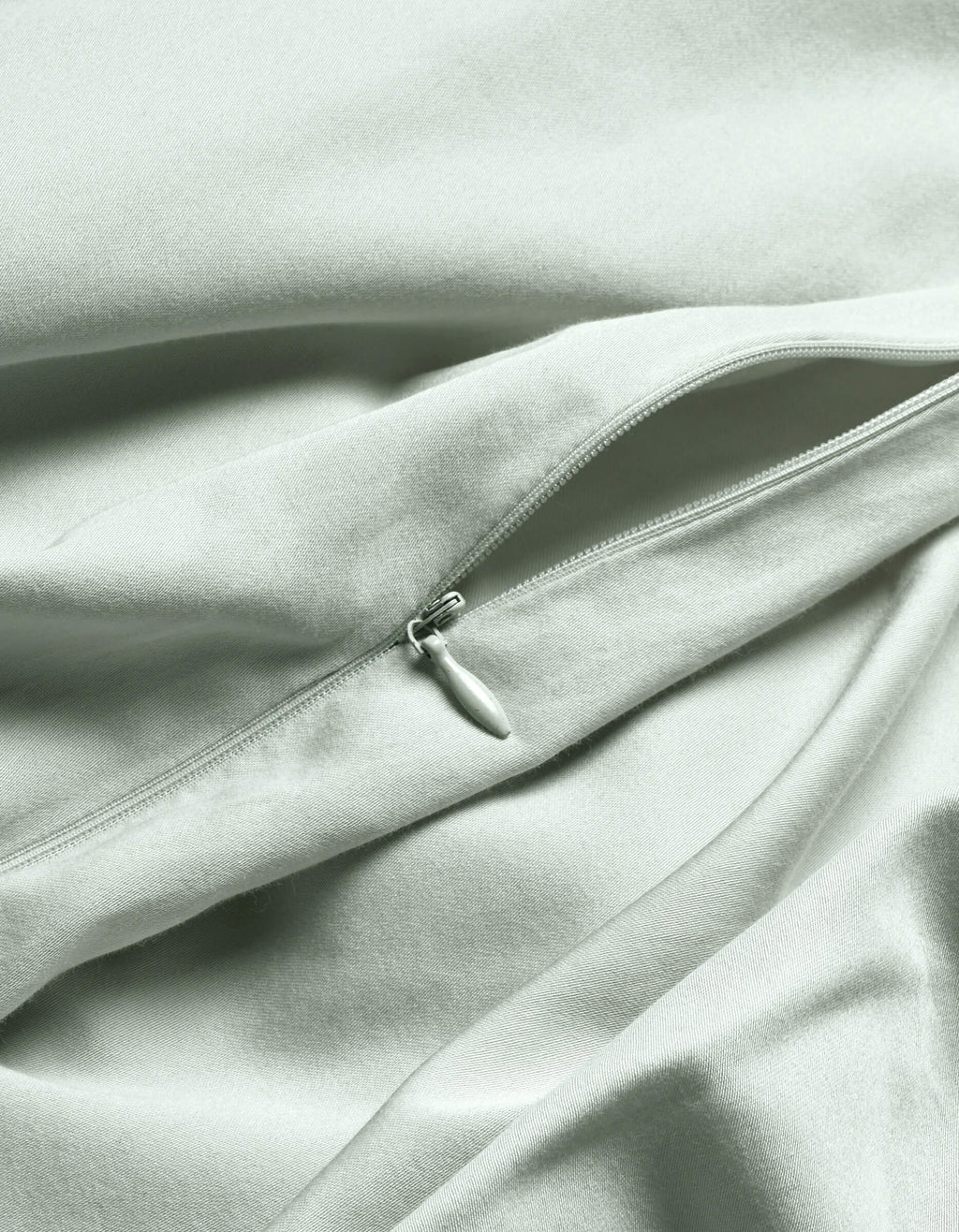 A close-up of the Cozy Earth Soft-Wash Cotton Duvet Cover in light gray or pale green shows a visible, partially unzipped zipper creating gentle folds and soft shadows on the smooth fabric.|Color:Laurel