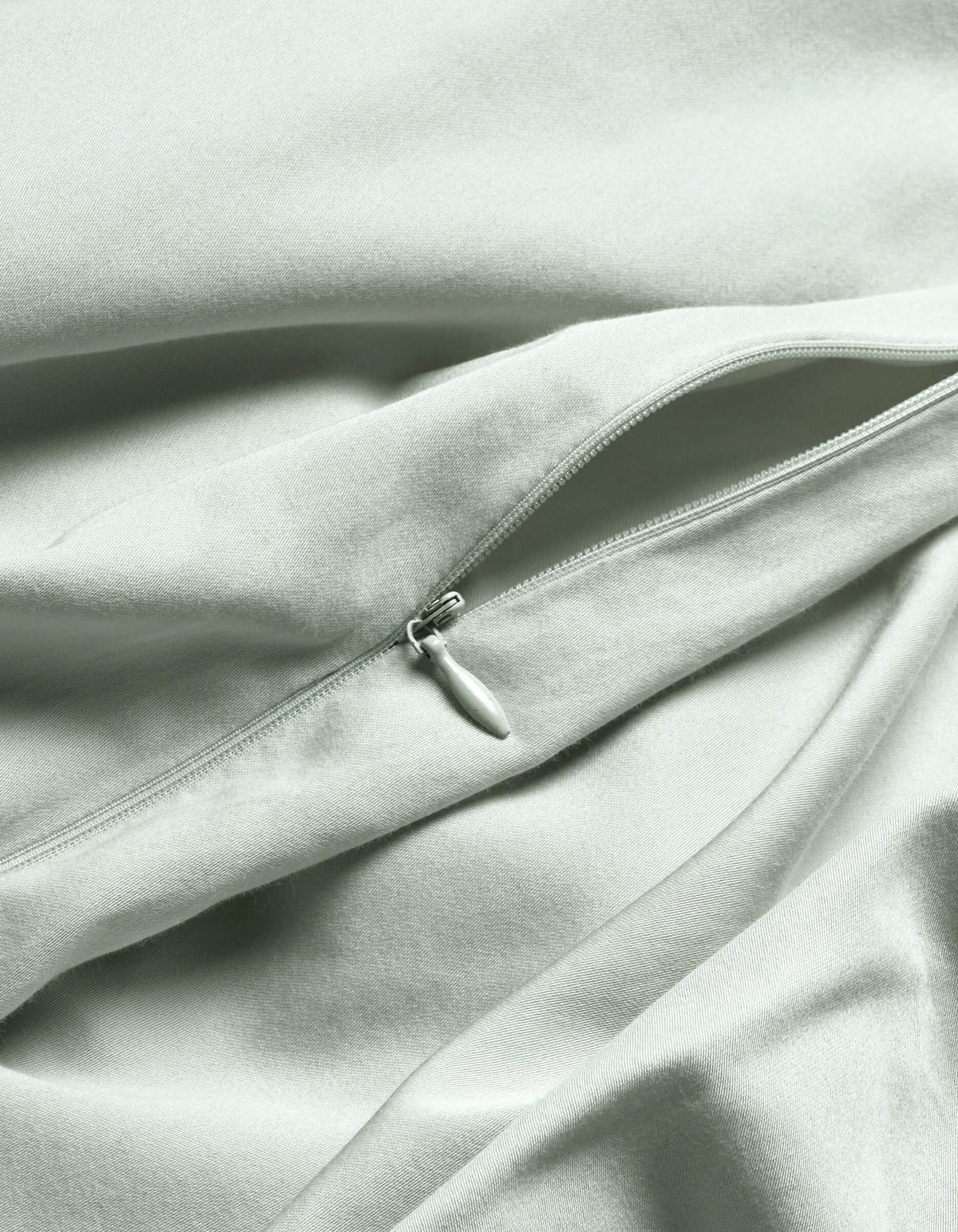 A close-up of the Cozy Earth Soft-Wash Cotton Duvet Cover in light gray or pale green shows a visible, partially unzipped zipper creating gentle folds and soft shadows on the smooth fabric.|Color:Laurel