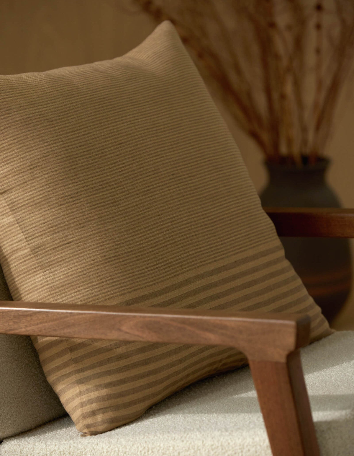 A close-up of a wooden armchair featuring the Cozy Earth Striped Throw Pillow, with a blurred vase of dried branches in the background.|Color:Mixed Stripe Sable and Sandalwood |Size:24" x 24"
