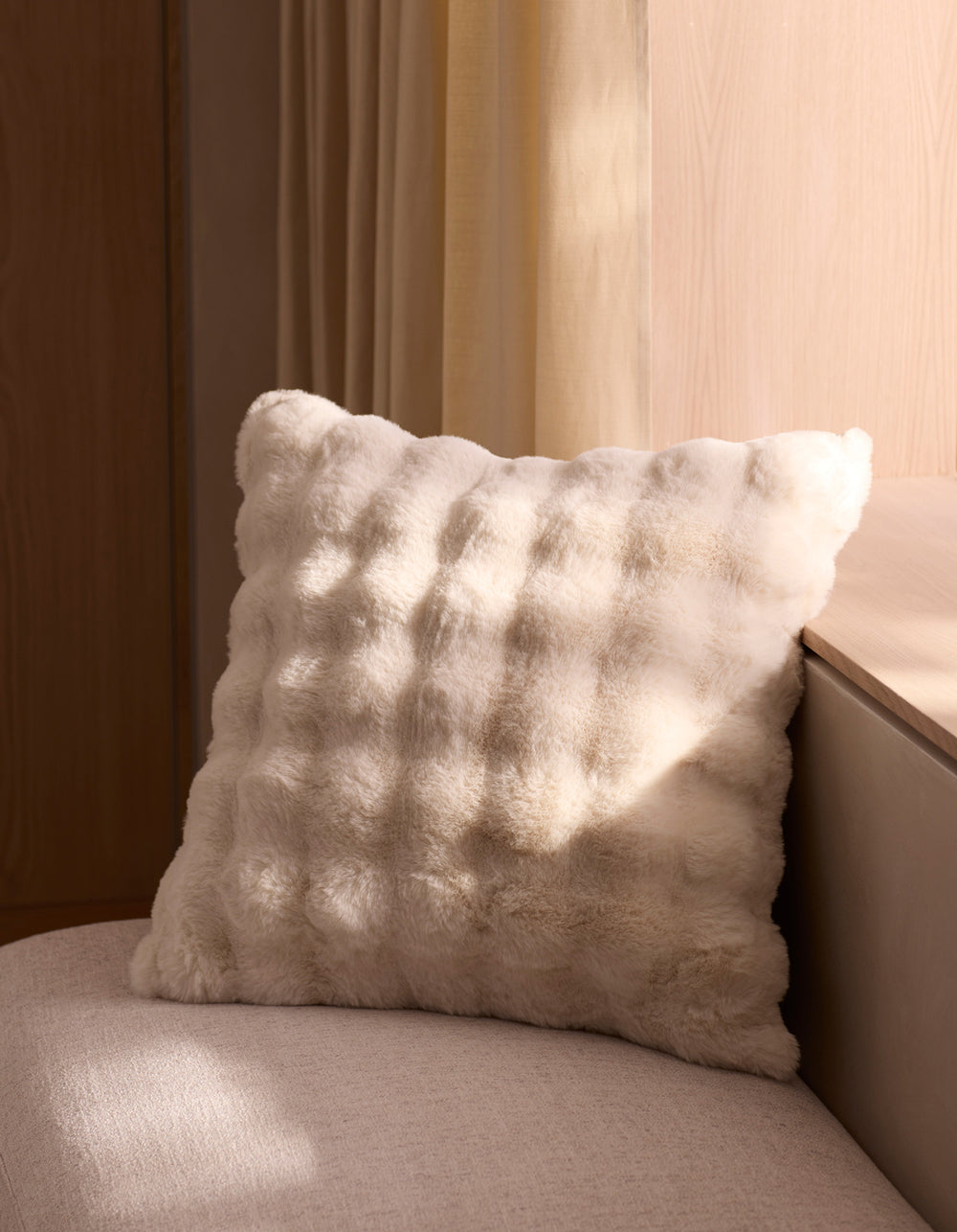 The Cozy Earth Bubble Cuddle Pillow, soft and fluffy in white, sits on a light beige seat near a wall as sunlight streams through the window, casting gentle shadows on the pillow and surrounding surfaces.|Color:Creme |Size:20" x 20"