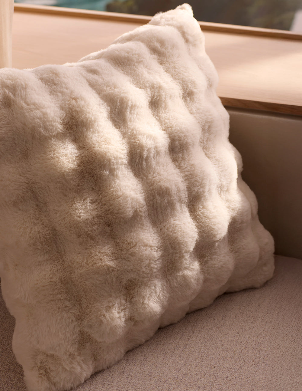 The Cozy Earth Bubble Cuddle Pillow, a soft cream faux fur with a raised texture, rests on a light beige couch by a wooden window ledge, illuminated by natural sunlight.|Color:Creme |Size:20" x 20"