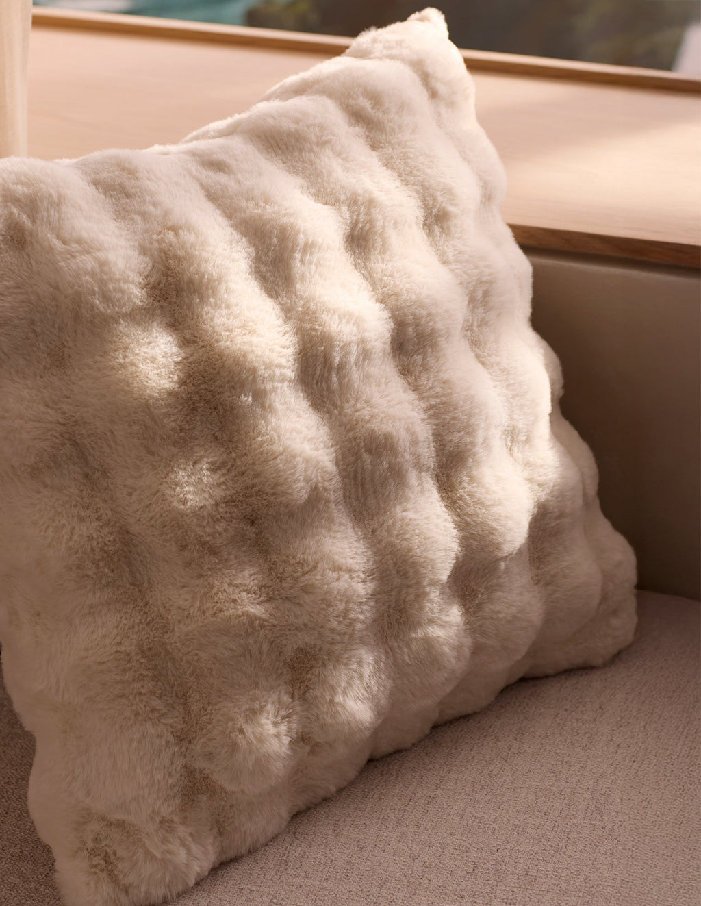 The Cozy Earth Bubble Cuddle Pillow, a soft cream faux fur with a raised texture, rests on a light beige couch by a wooden window ledge, illuminated by natural sunlight.|Color:Creme |Size:24" x 24"