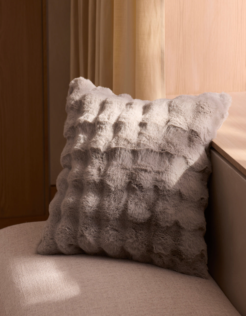 The Bubble Cuddle Pillow by Cozy Earth, featuring textured gray faux fur, rests on a light beige upholstered bench beside a wooden wall and cream curtains, with natural light casting gentle shadows.|Color:Light Grey |Size:20" x 20"