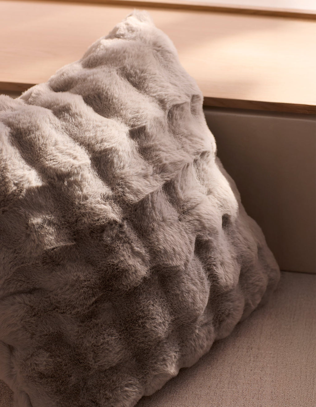 The Bubble Cuddle Pillow by Cozy Earth, featuring soft gray faux fur with a wavy texture, rests on a light wooden surface in a sunlit, neutral-toned room.|Color:Light Grey |Size:24" x 24"