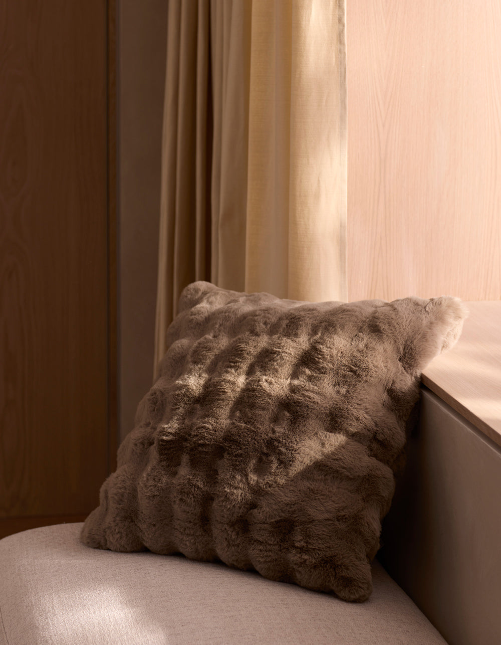 The Cozy Earth Bubble Cuddle Pillow sits on a light beige cushioned bench by pale wood walls and a window with cream curtains, softly illuminated by natural light.|Color:Walnut |Size:20" x 20"