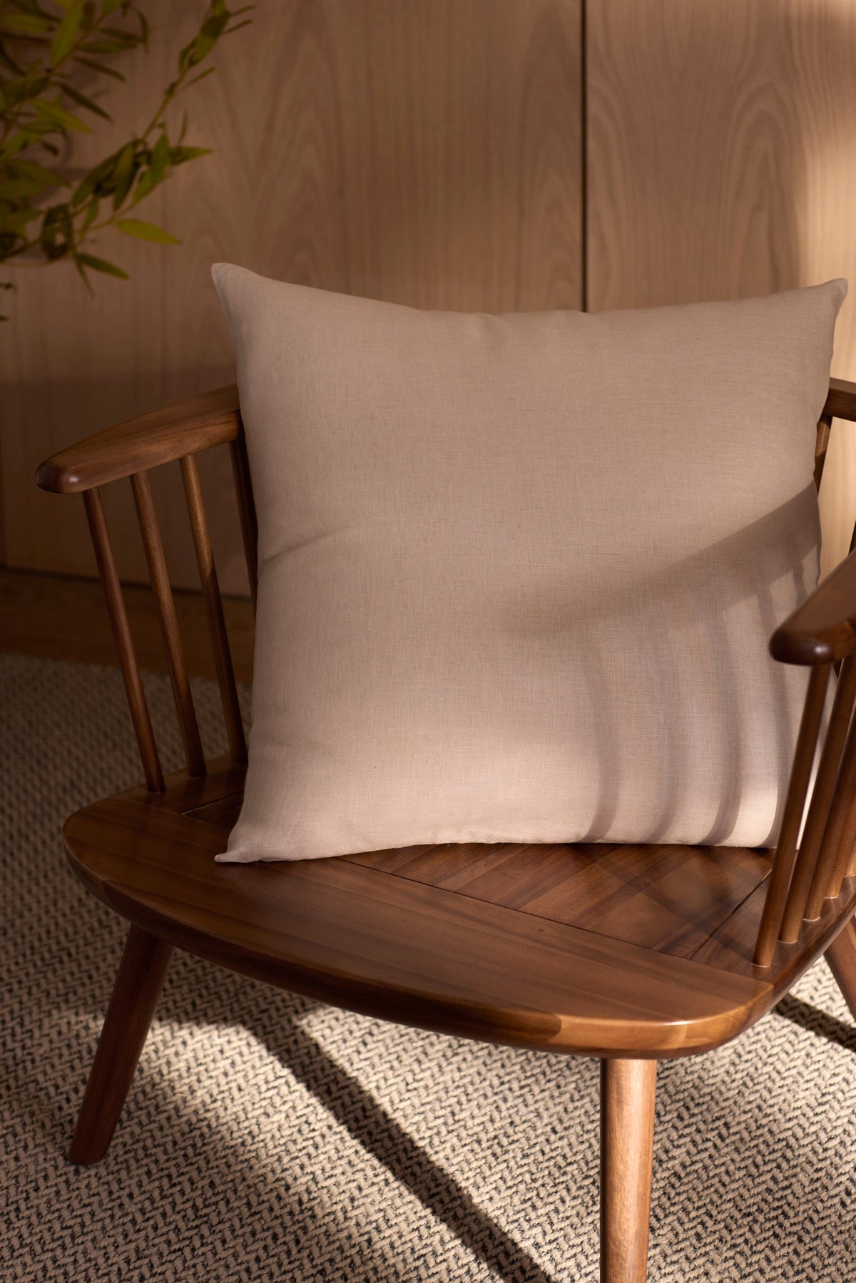 A Cozy Earth Linen Throw Pillow rests on a wooden chair with a light beige cushion atop a textured woven rug, as sunlight casts soft shadows and a green plant peeks in the background. |Color:Birch |Size:24" x 24"